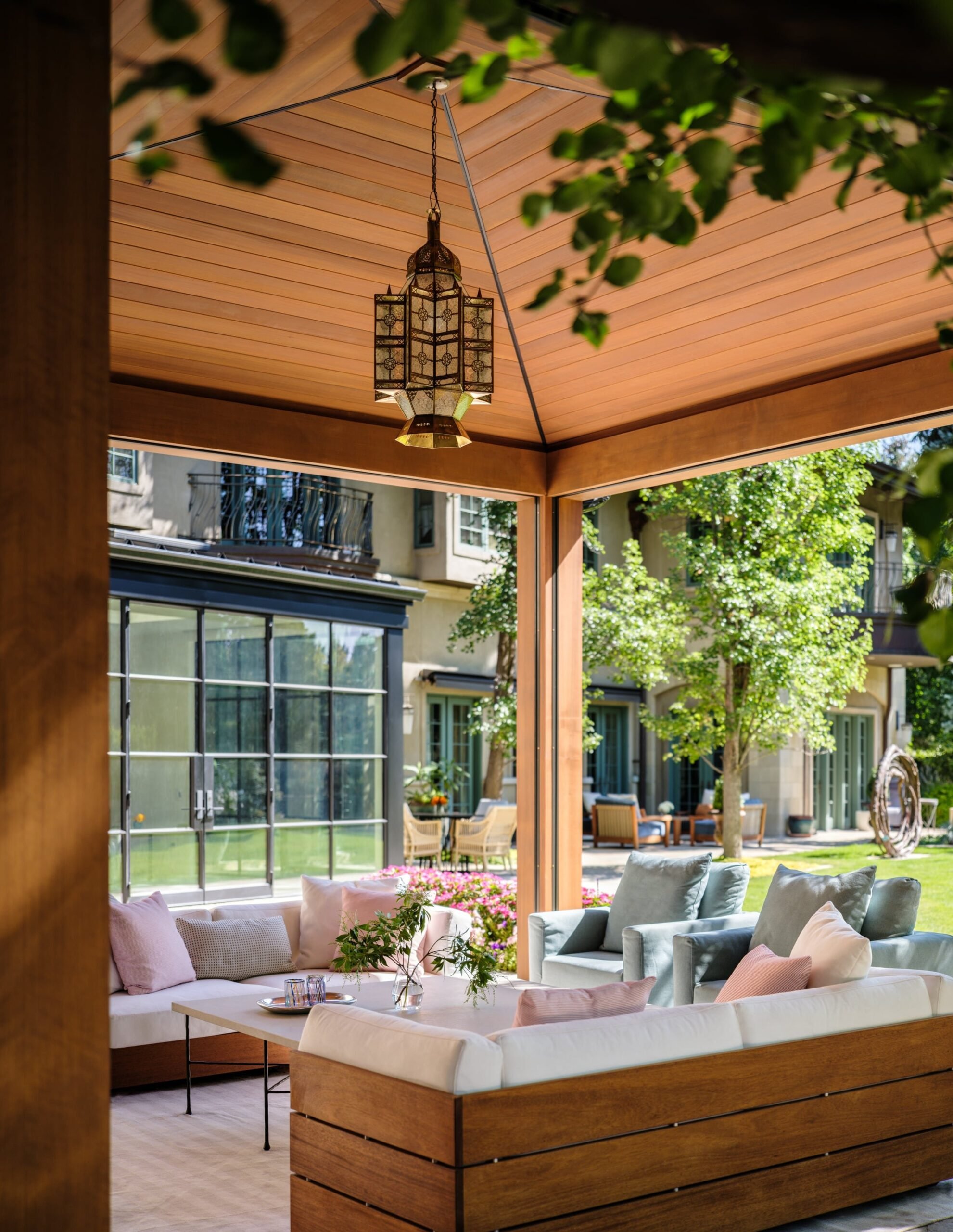 View of an indoor-outdoor living space with a seating area featuring a white sectional sofa and pastel-colored pillows, overlooking a garden with trees, flowers, and outdoor seating, with a wooden ceiling and a hanging ornate lantern. Bonacina. 