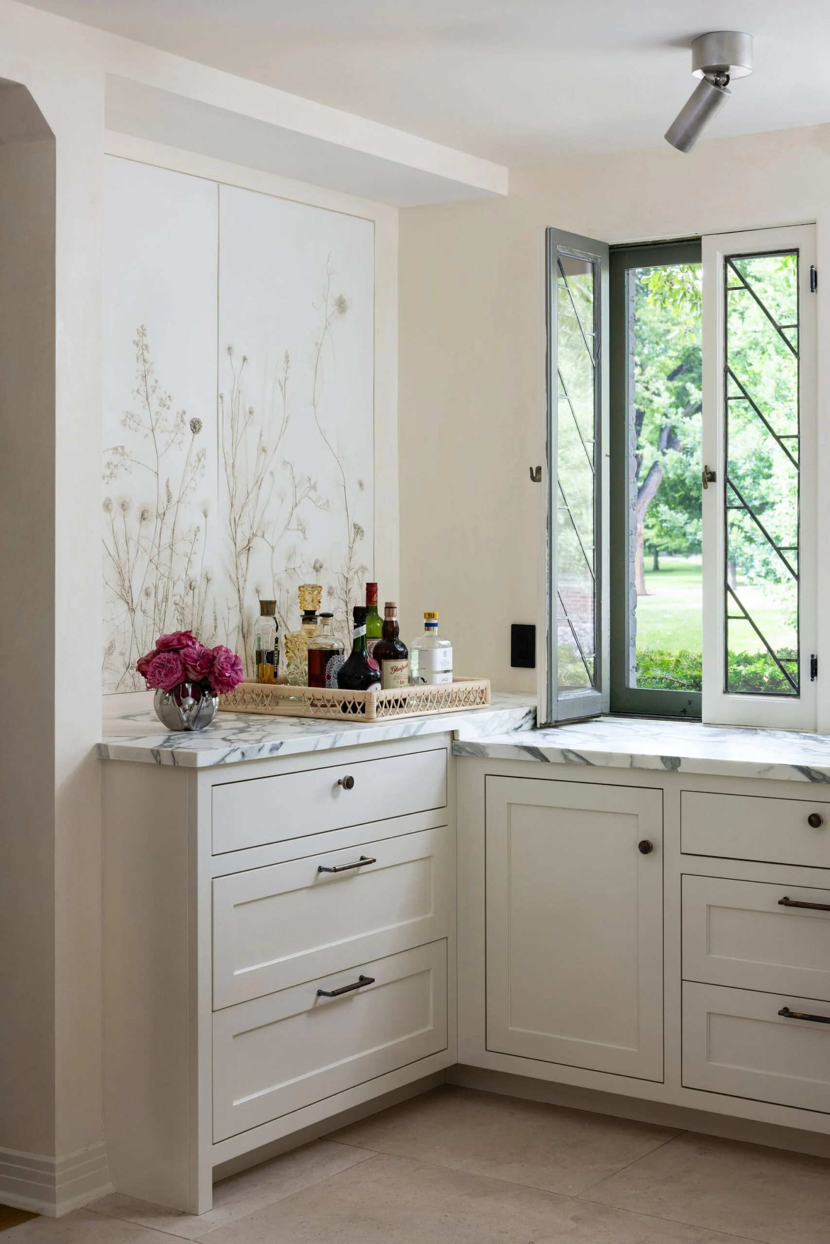 Kitchen corner with white cabinets, marble countertops, and open window showing green outdoor scenery, with a tray of liquor bottles and a vase of pink roses on the counter. Charleston interior designer. 