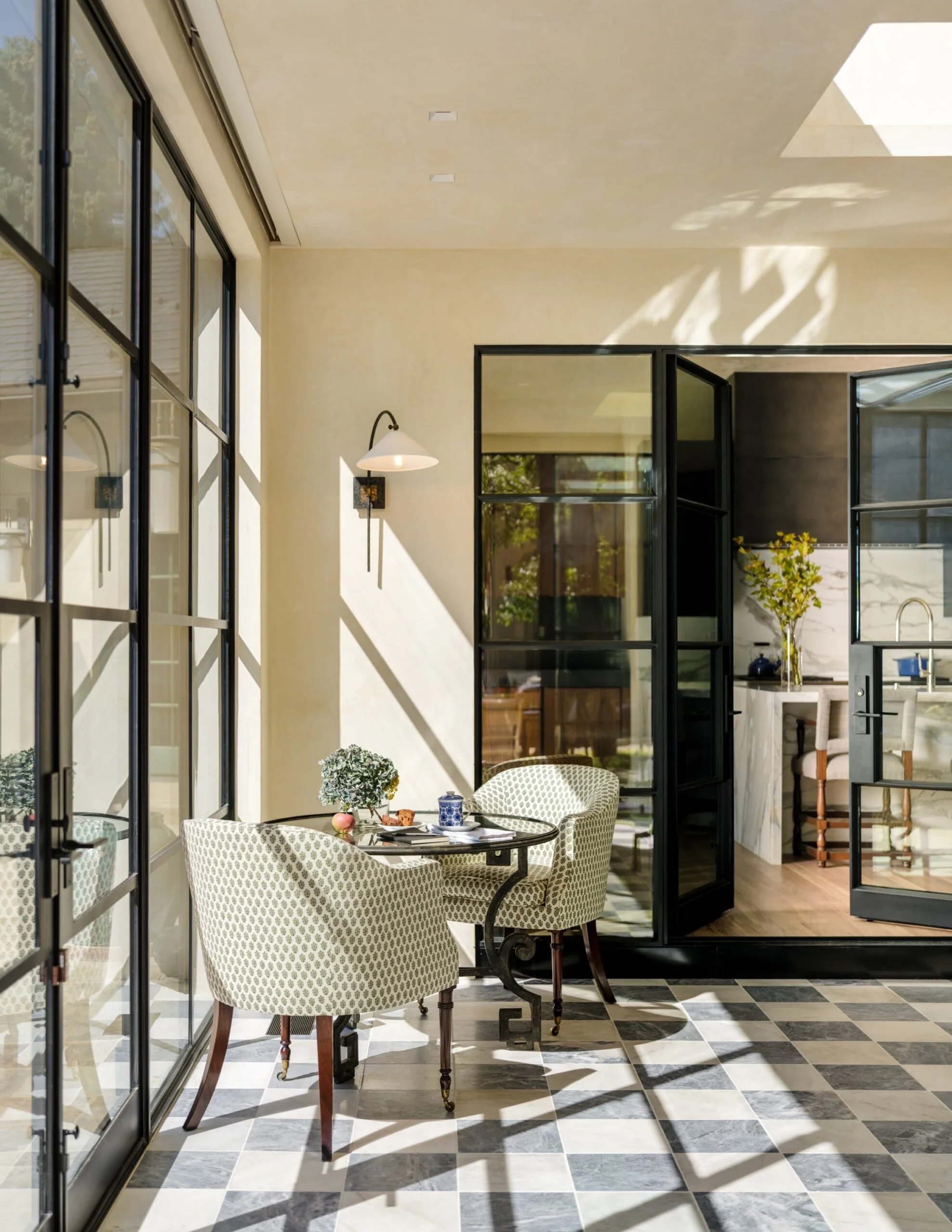 Bright sunlit dining area with checkered black and white floor tiles, a small round table, two patterned chairs, and a black-framed glass wall with a view into the kitchen. Charleston interior designer Sarah Hart Interior Design. Jamb London chairs. 