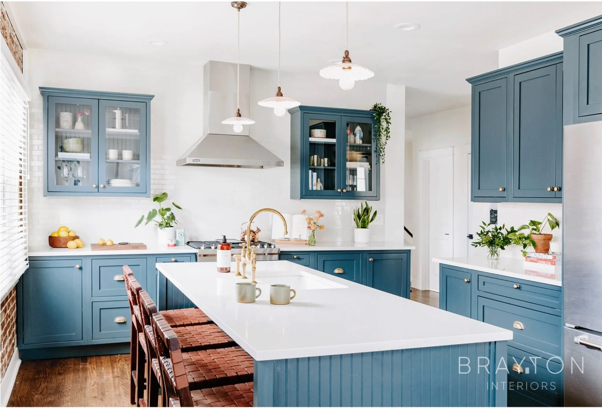 Modern kitchen with blue cabinets, white countertops, and a large island. Copper pendant lights hang above the island. There are plants, a bowl of lemons, and kitchen utensils on the counters. A stainless steel range hood and refrigerator are visible