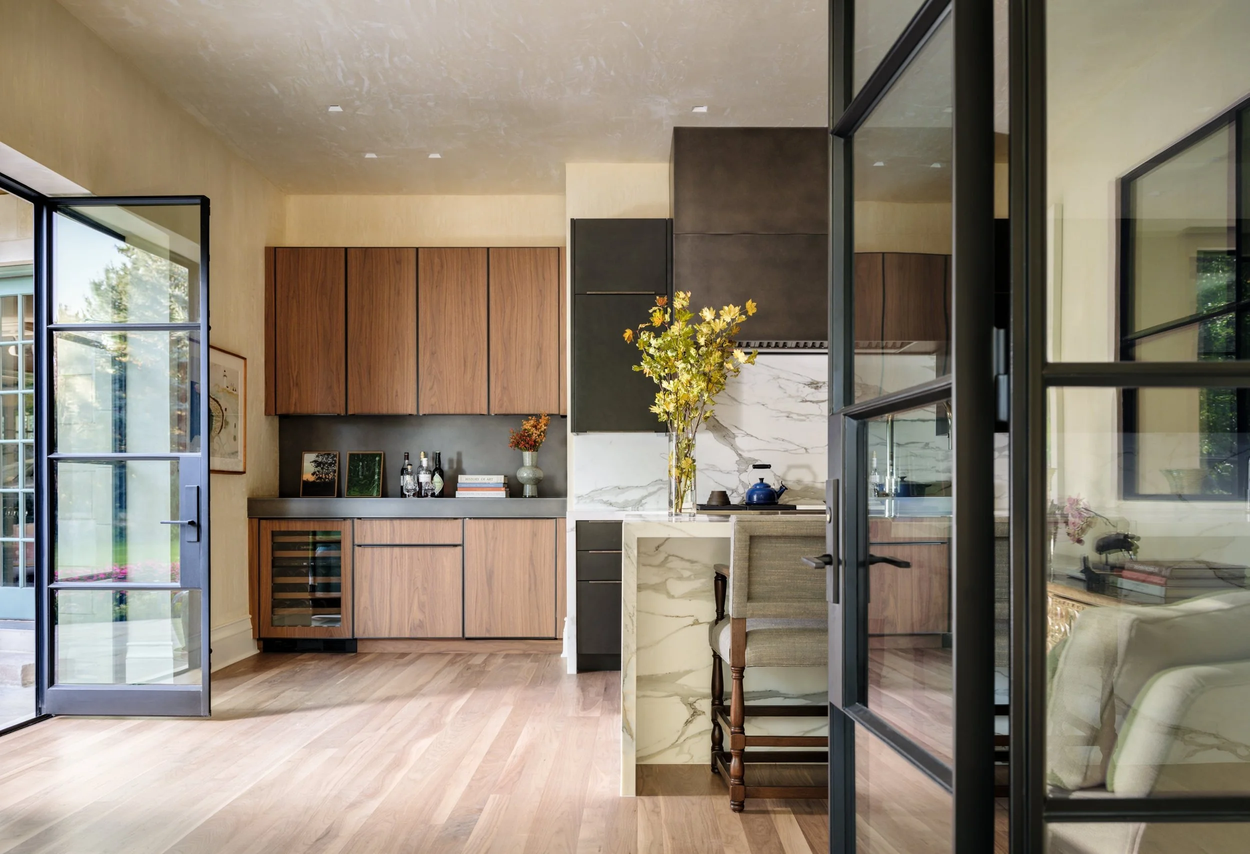 Modern kitchen with wooden cabinets, marble countertops, a small bar area with a chair, a large blue teapot, and glass doors leading outside. Charleston interior designer Sarah Hart Interior Design. 