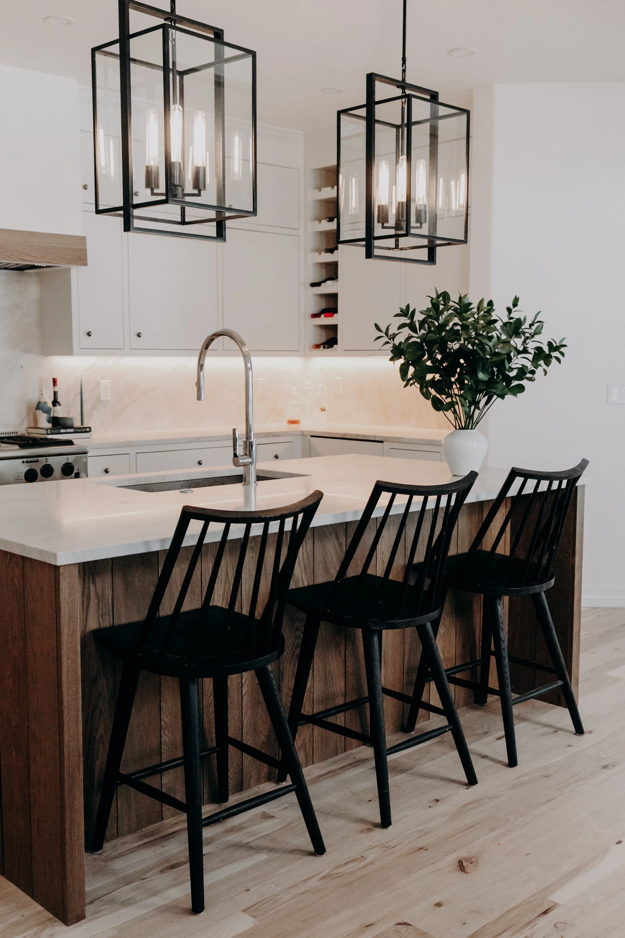 Modern kitchen with white cabinets, marble countertops, black bar stools, and a large potted plant on the island under two black pendant lights.