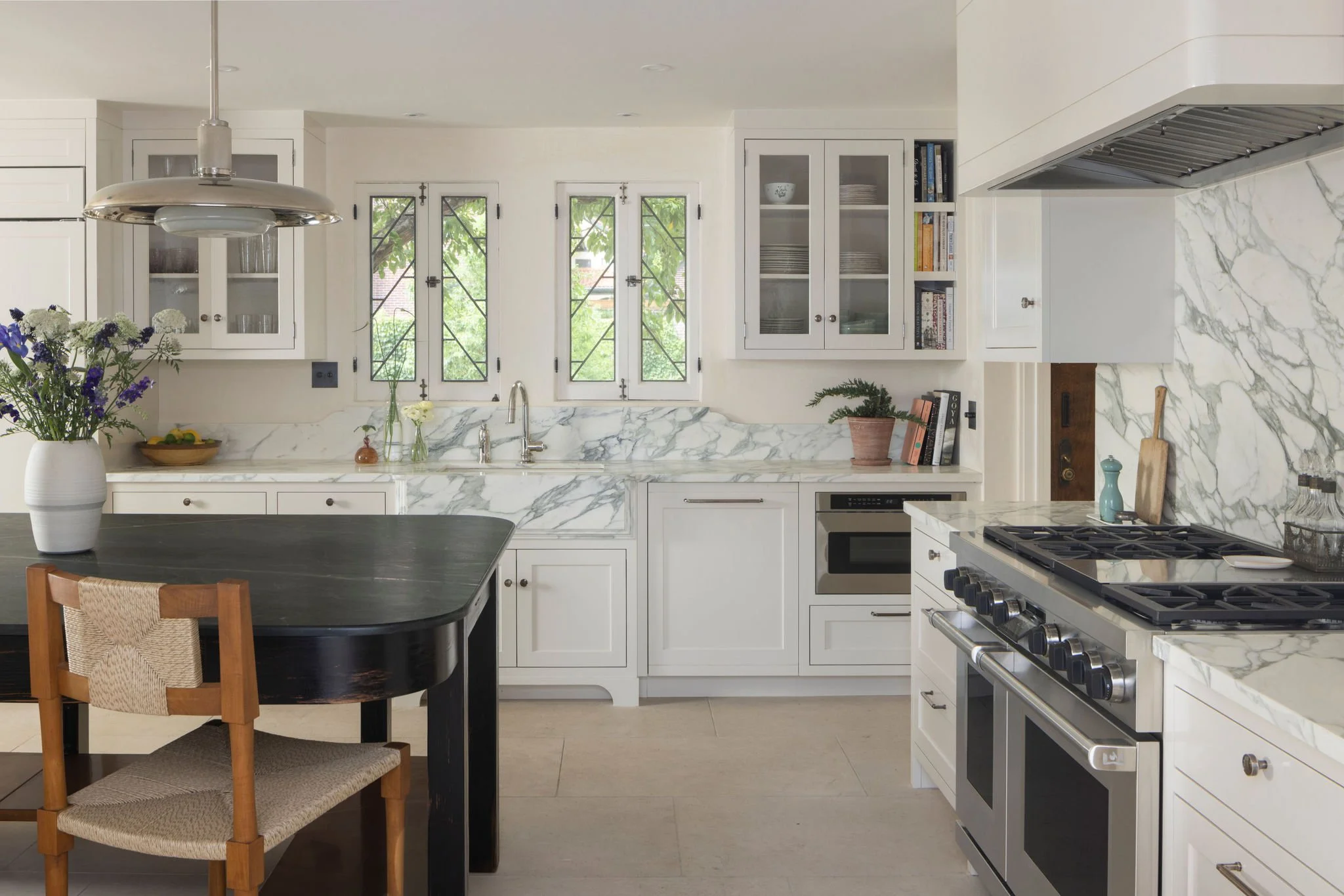 Modern kitchen with white cabinets, marble countertops, and a black dining table with a chair. Decorative plants and flowers are on the counters and table. Hollywood at Home barstools and Roman and Williams pendants. Waterworks plumbing. 