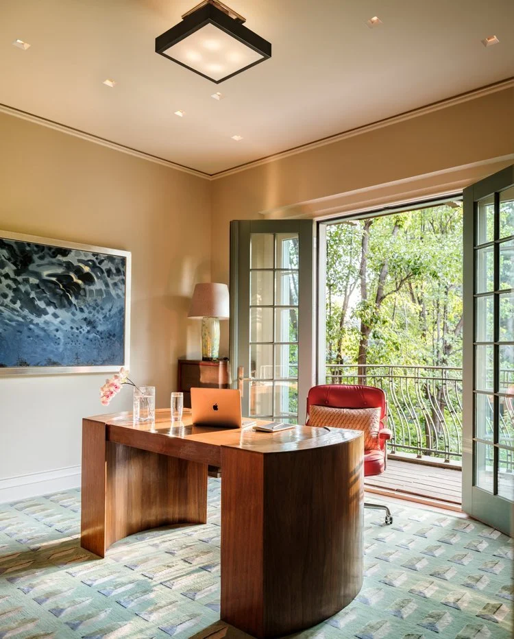 Home office with wooden desk, laptop, glass of water, red armchair, large window opening to a green tree-filled balcony. Rose Tarlow desk, Holland and Sherry Rug. Urban Electric lighting. Juin Ho cabinets. Charleston Interior designer. 