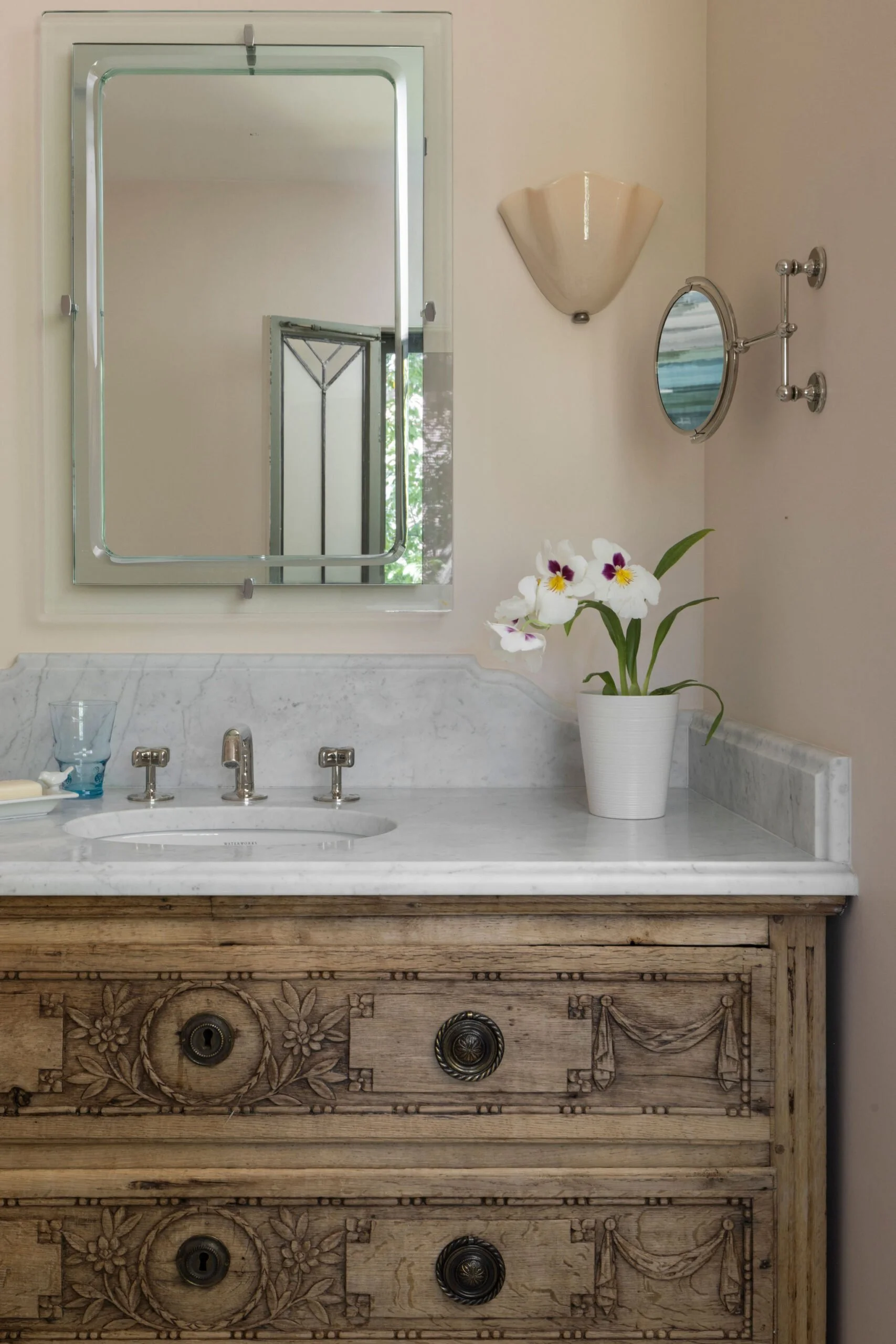 A bathroom vanity with a marble top, a mirror with a metal frame, flowers in a white pot, a wall sconce, and a small round magnifying mirror on a metallic arm. Interior design Charleston. 