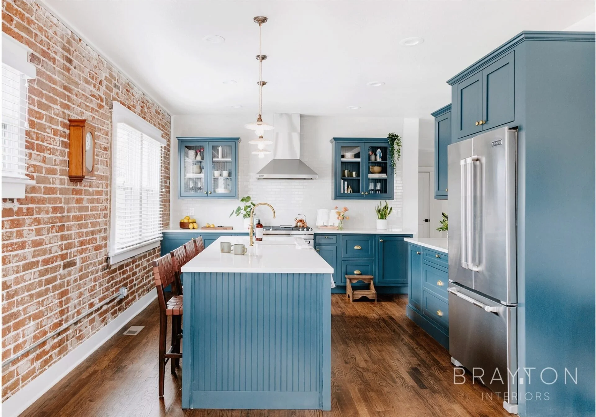 Modern kitchen with blue cabinets, white countertops, and a brick accent wall. Features include a large kitchen island, stainless steel refrigerator, and decorative pendant lights.
