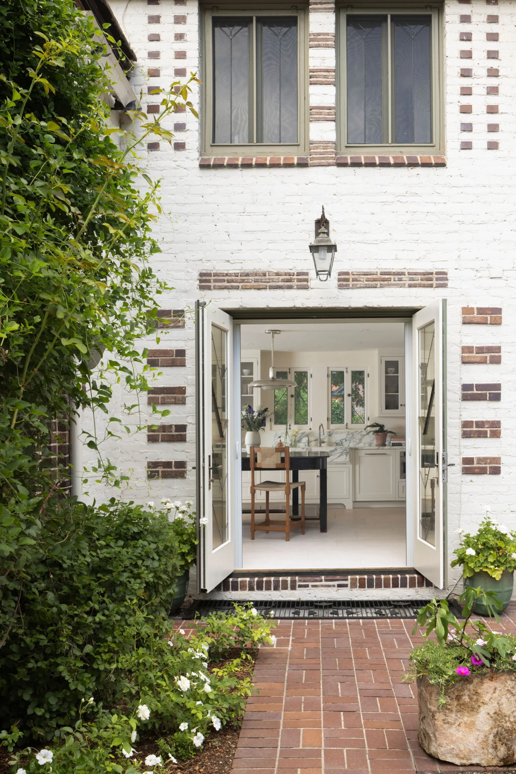 Open French doors leading into a modern kitchen from a brick patio area, with brick and white painted brick exterior, flowering plants, and shrubs outside. Historic home renovation by Charleston Interior Designer Sarah Hart .