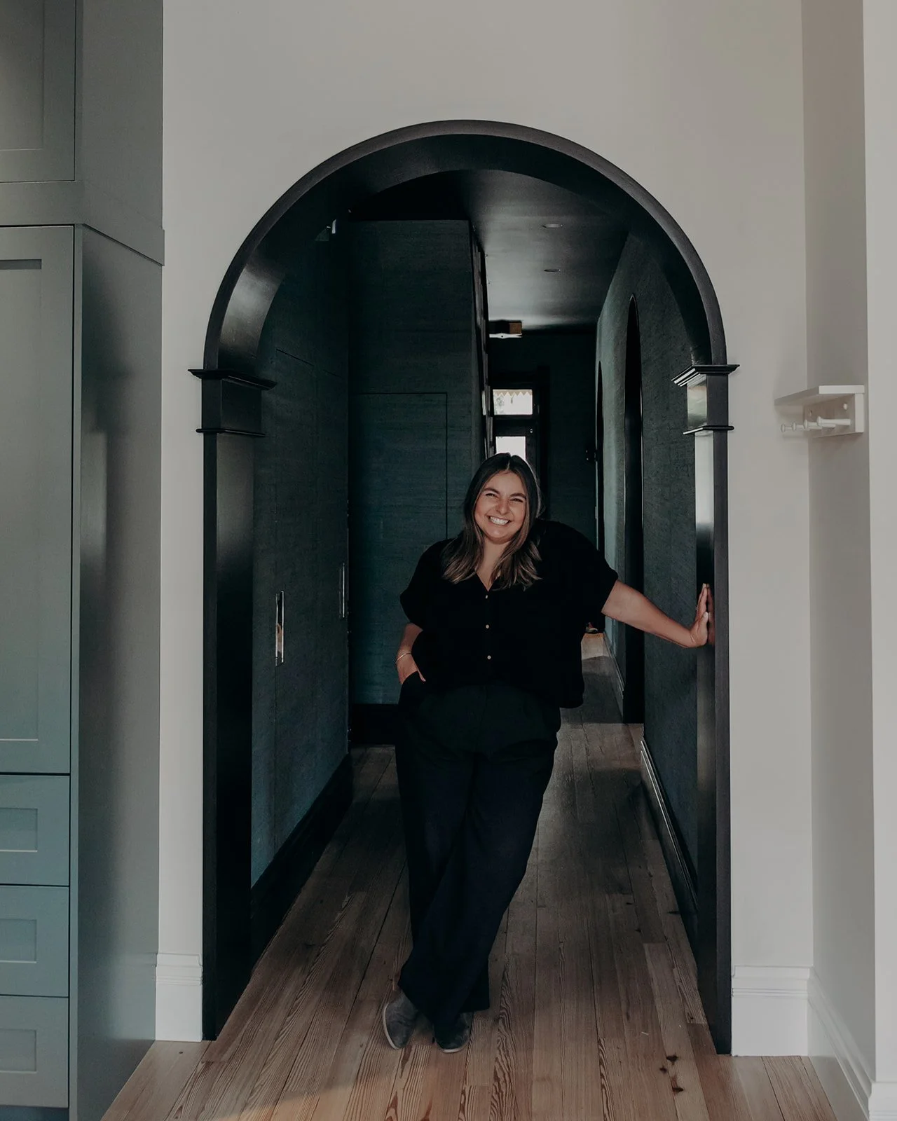 A woman standing in a hallway on a wooden floor, smiling with her hand on a wall, with dark walls and an archway behind her.