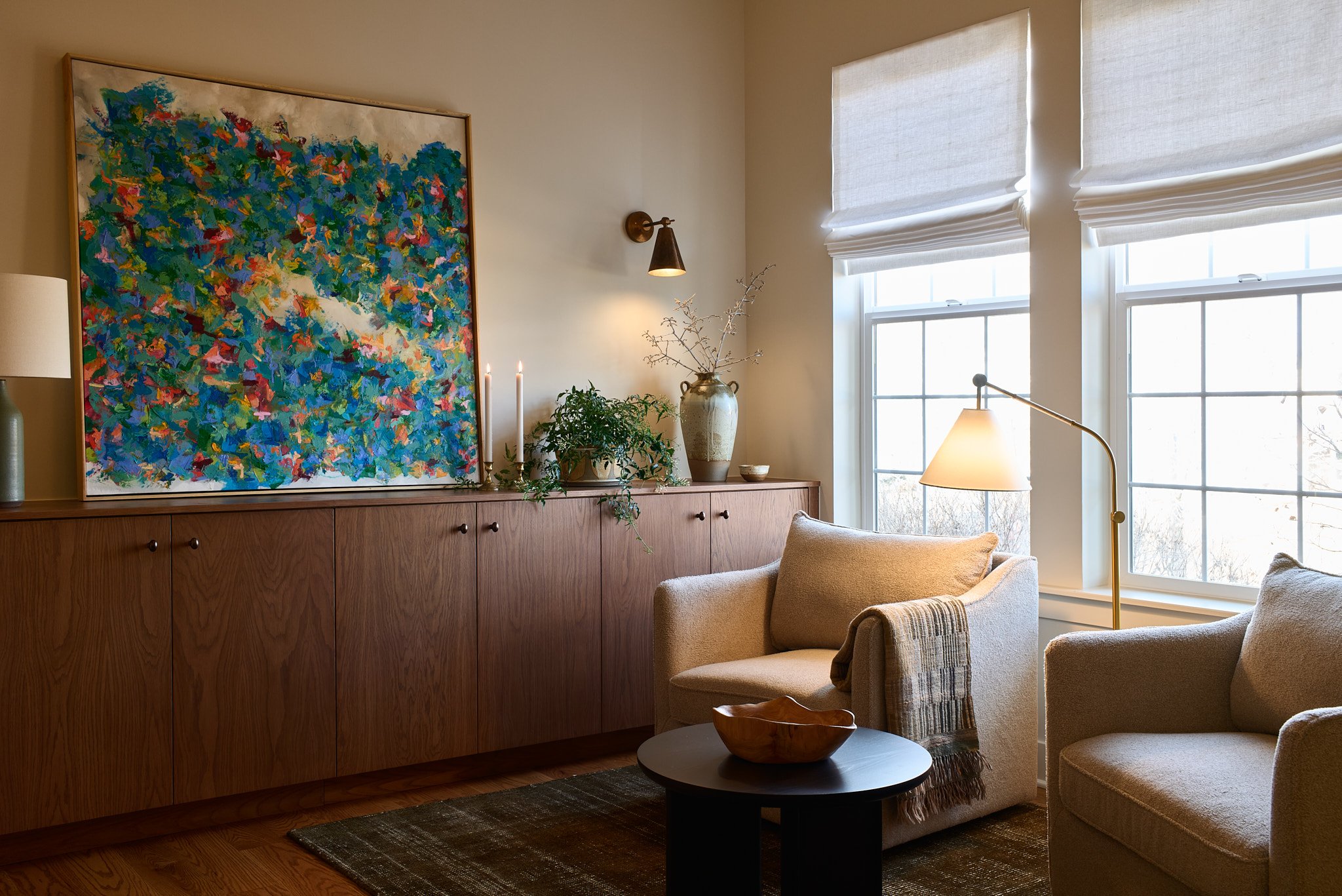 Living room with beige armchair, black round coffee table with a wooden bowl, and a sofa. Large windows with white blinds, a wooden console with plants, candles, and artwork. Soft lighting and warm decor.