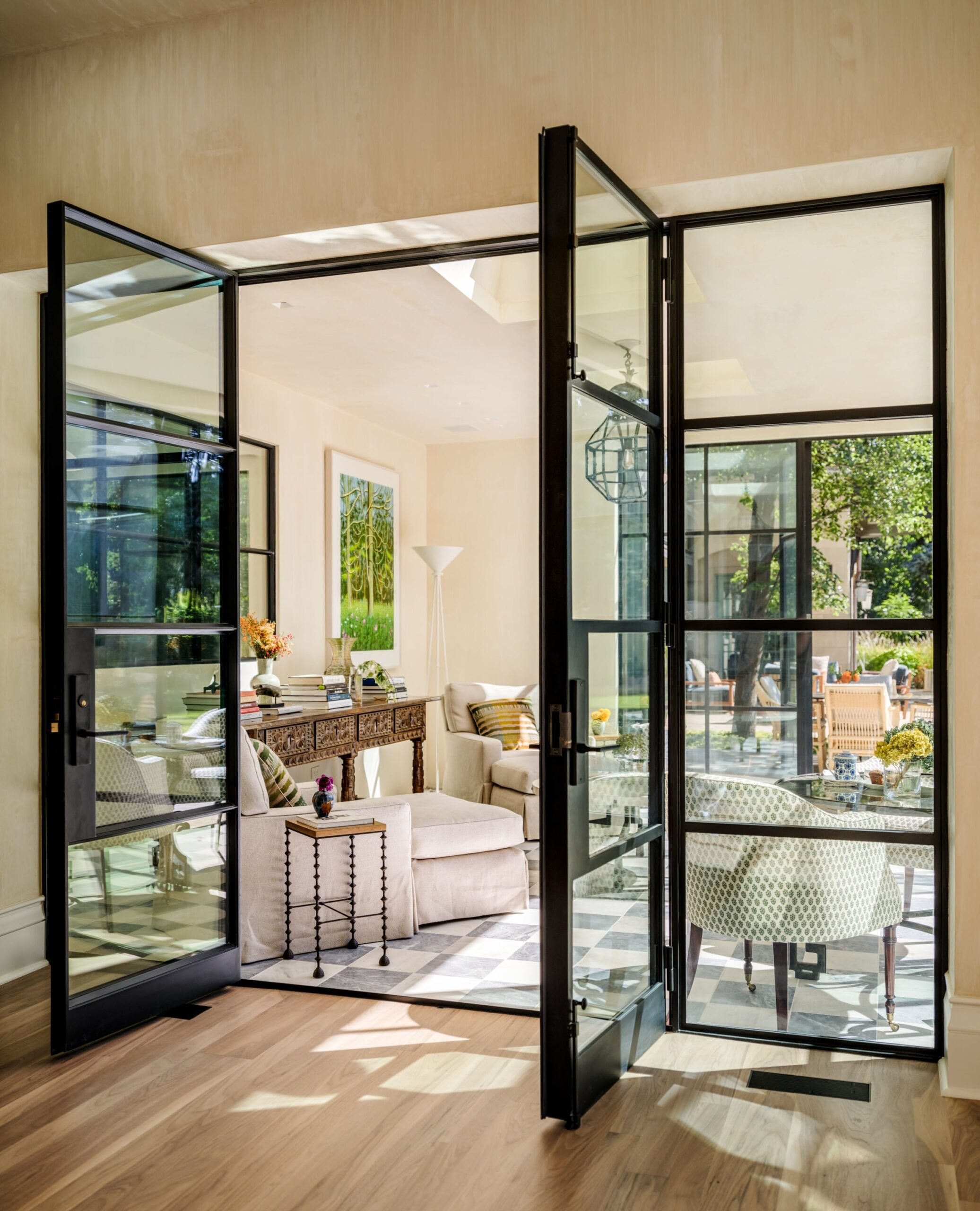 Interior view of a living room seen through black-framed glass doors, showing a sofa, armchair, artwork, and decor, with outdoor patio and greenery visible outside.
