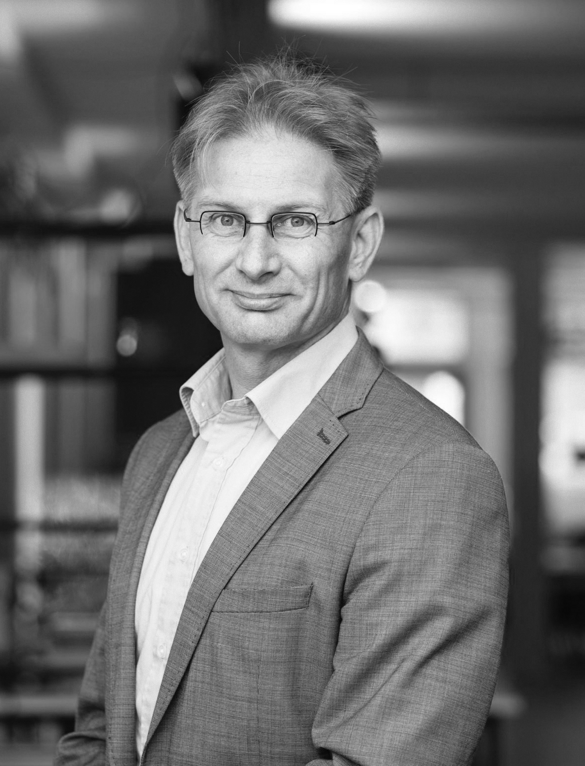 Zwart-wit portret van een man met grijs haar, bril en een pak, staand in een kantooromgeving.