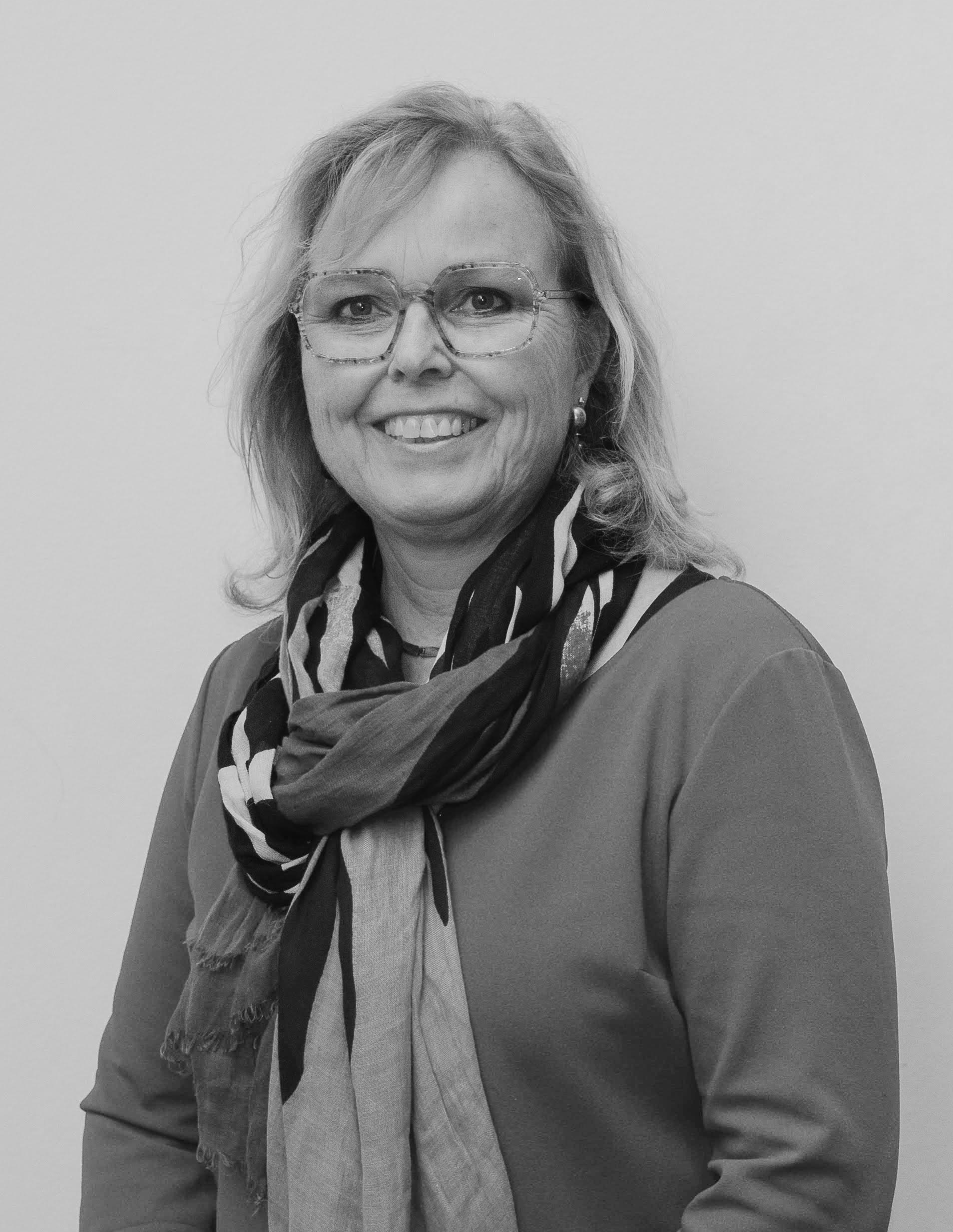 Een vrouw met lichtbruin haar, draagt een bril, een casual jas en een gestreepte scarf, lachend naar de camera.