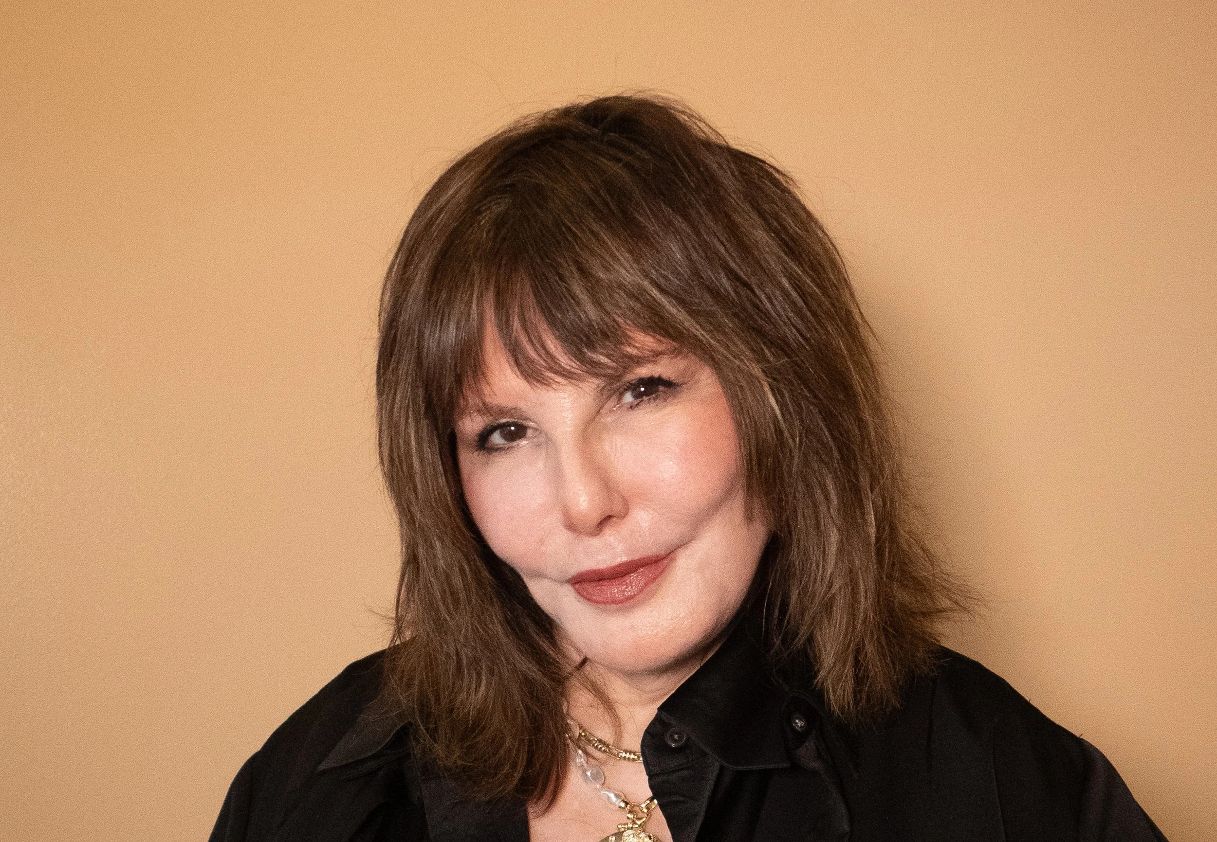 A woman with shoulder-length brown hair and bangs, wearing a black shirt and gold jewelry, smiling slightly and winking against a beige background.