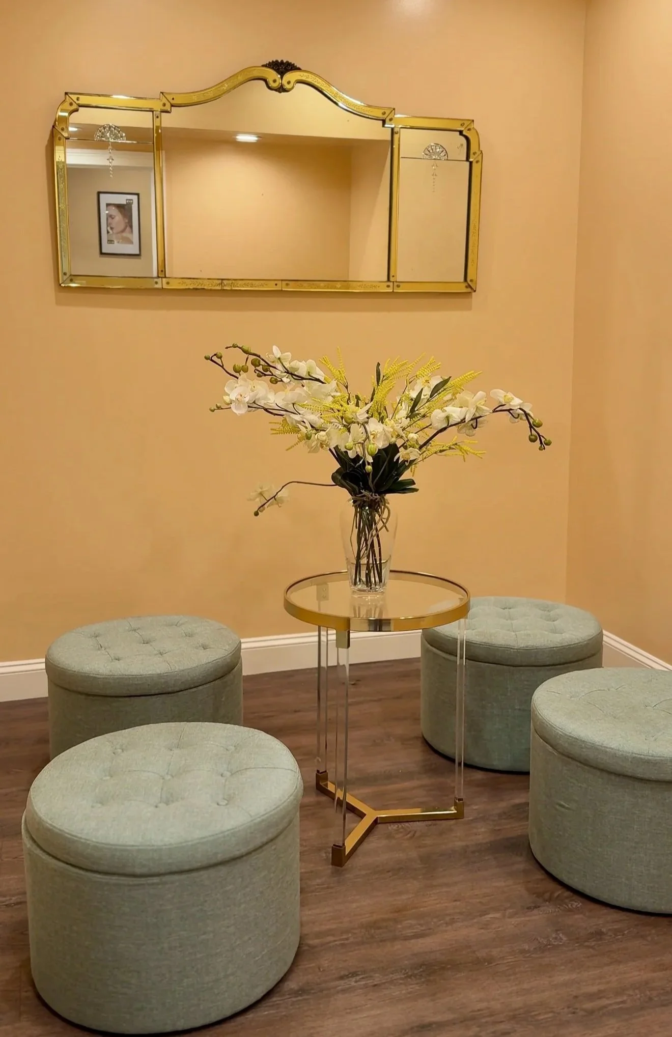A round glass-top table with a flower arrangement in a vase surrounded by four green upholstered ottomans in a room with beige walls, a gold-framed mirror, and wooden flooring.