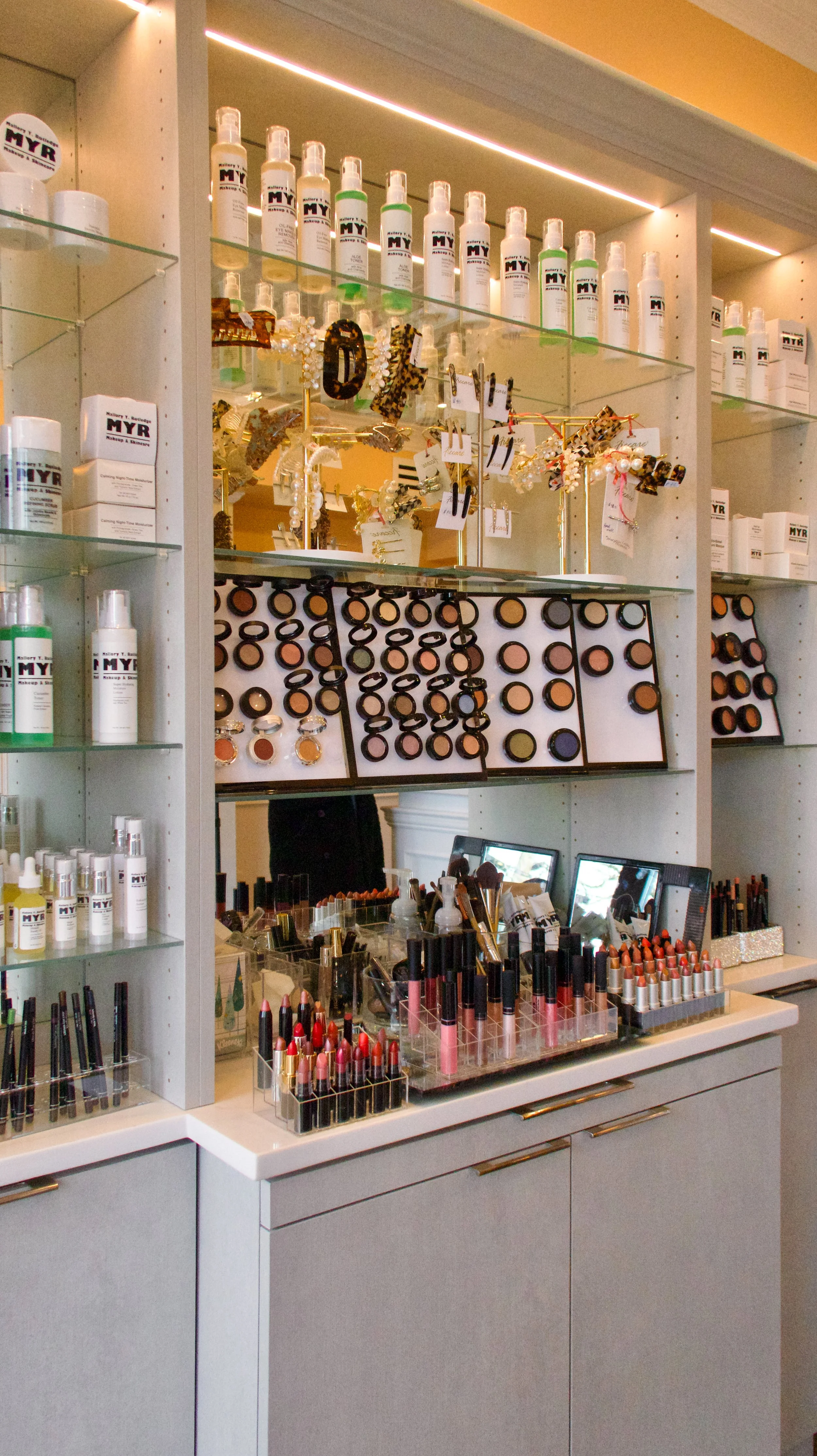 A makeup station with various cosmetics, brushes, and accessories on a countertop and glass shelves, including lipsticks, powders, spray bottles, and jewelry with leopard print accents.