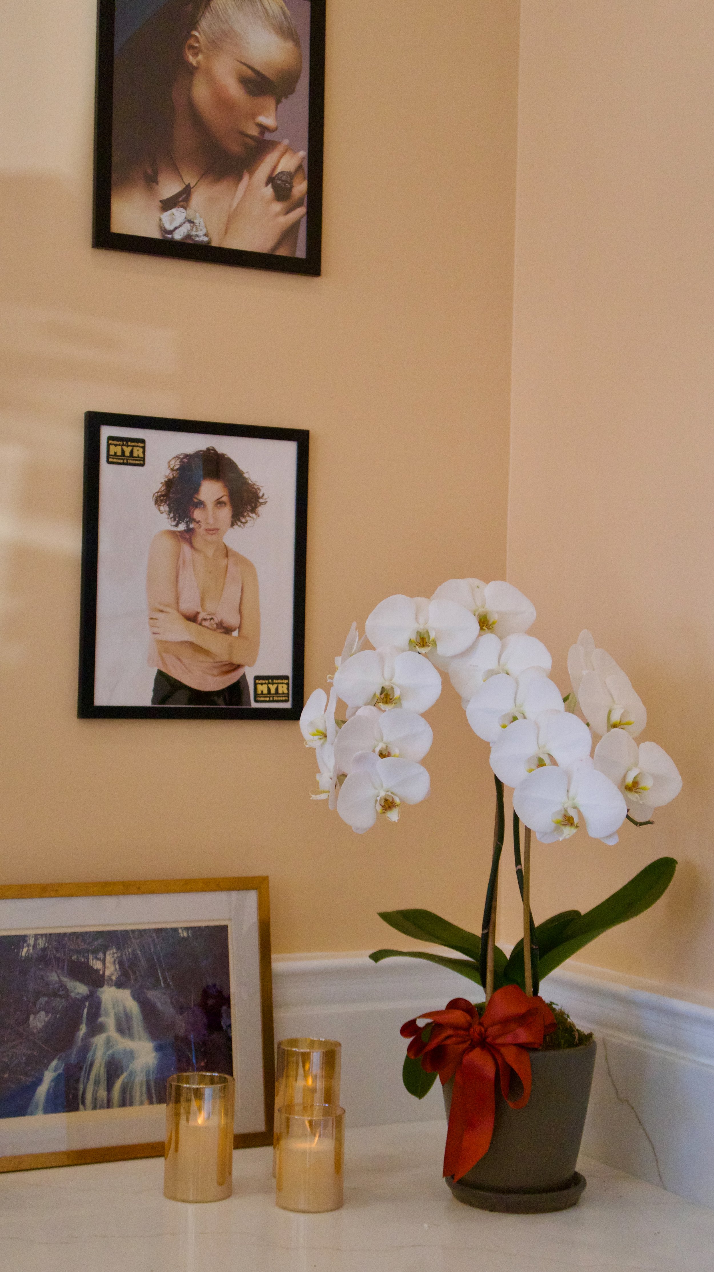 A potted white orchid with a red ribbon, framed waterfall photo, three gold candle holders, and framed photographs of women on a beige wall.