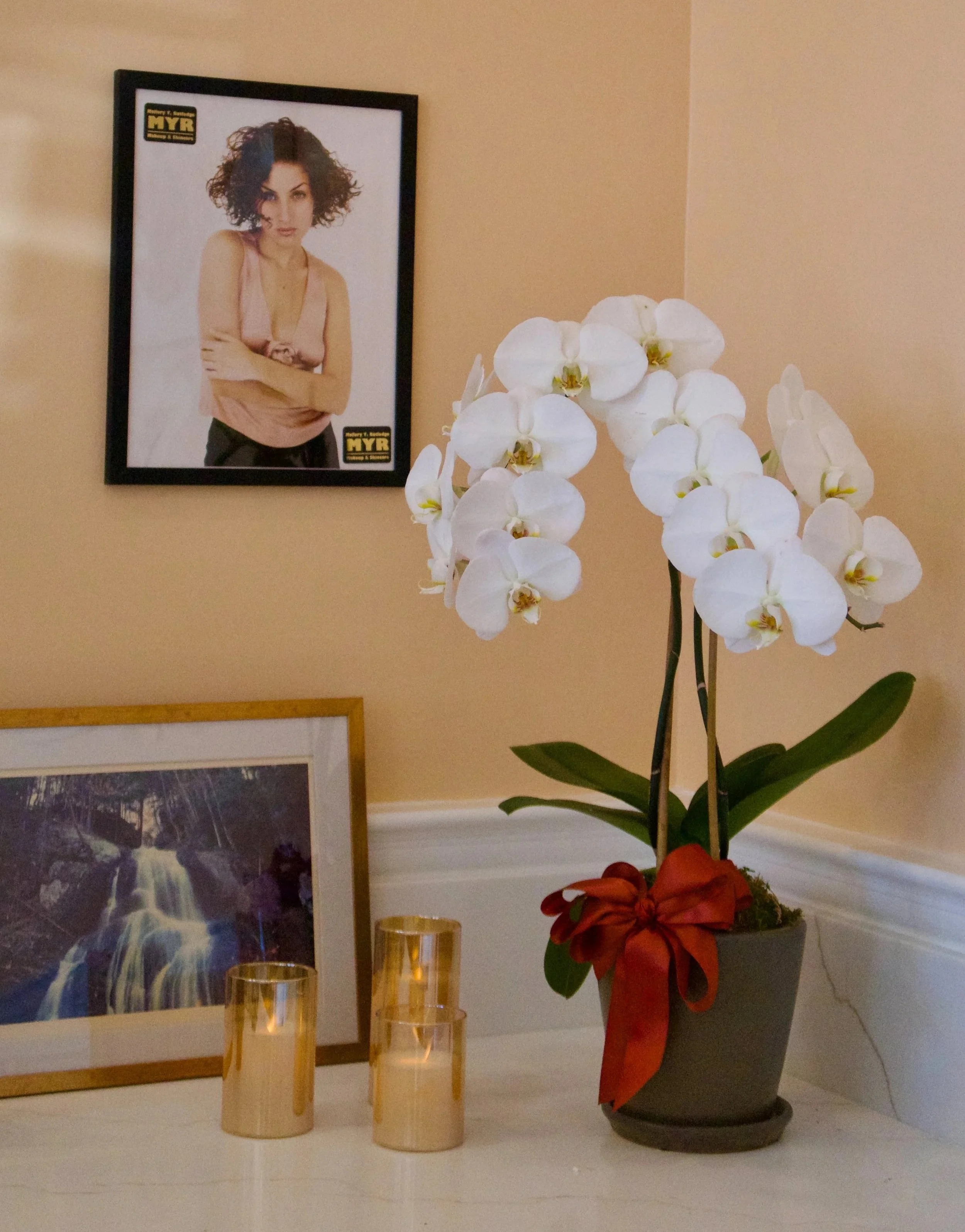 A white orchid plant with large blooms and green leaves in a gray pot decorated with a red bow, placed on a white surface. Behind it, a framed landscape photo of a waterfall leans against the wall, and three gold-colored candles are arranged next to it. On the wall above, there is a framed poster of a woman with curly hair.
