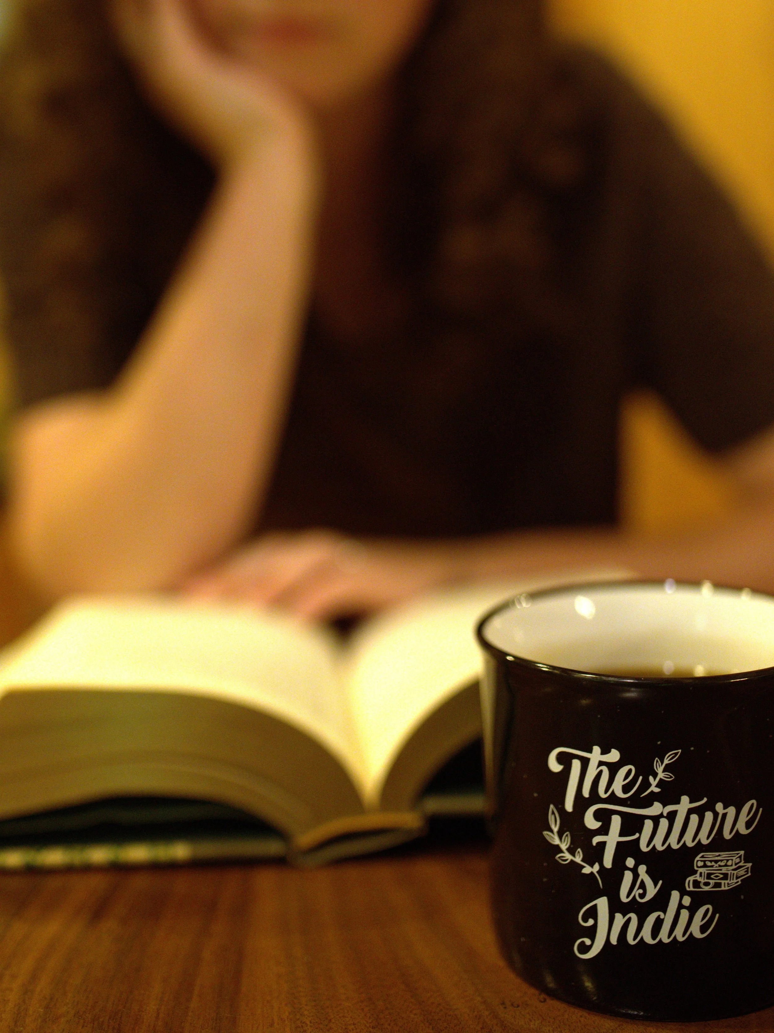 Close-up of a black mug with white text that reads 'The Future is Indie' next to an open book on a wooden surface. A woman is blurred in the background, resting her chin on her hand and reading.