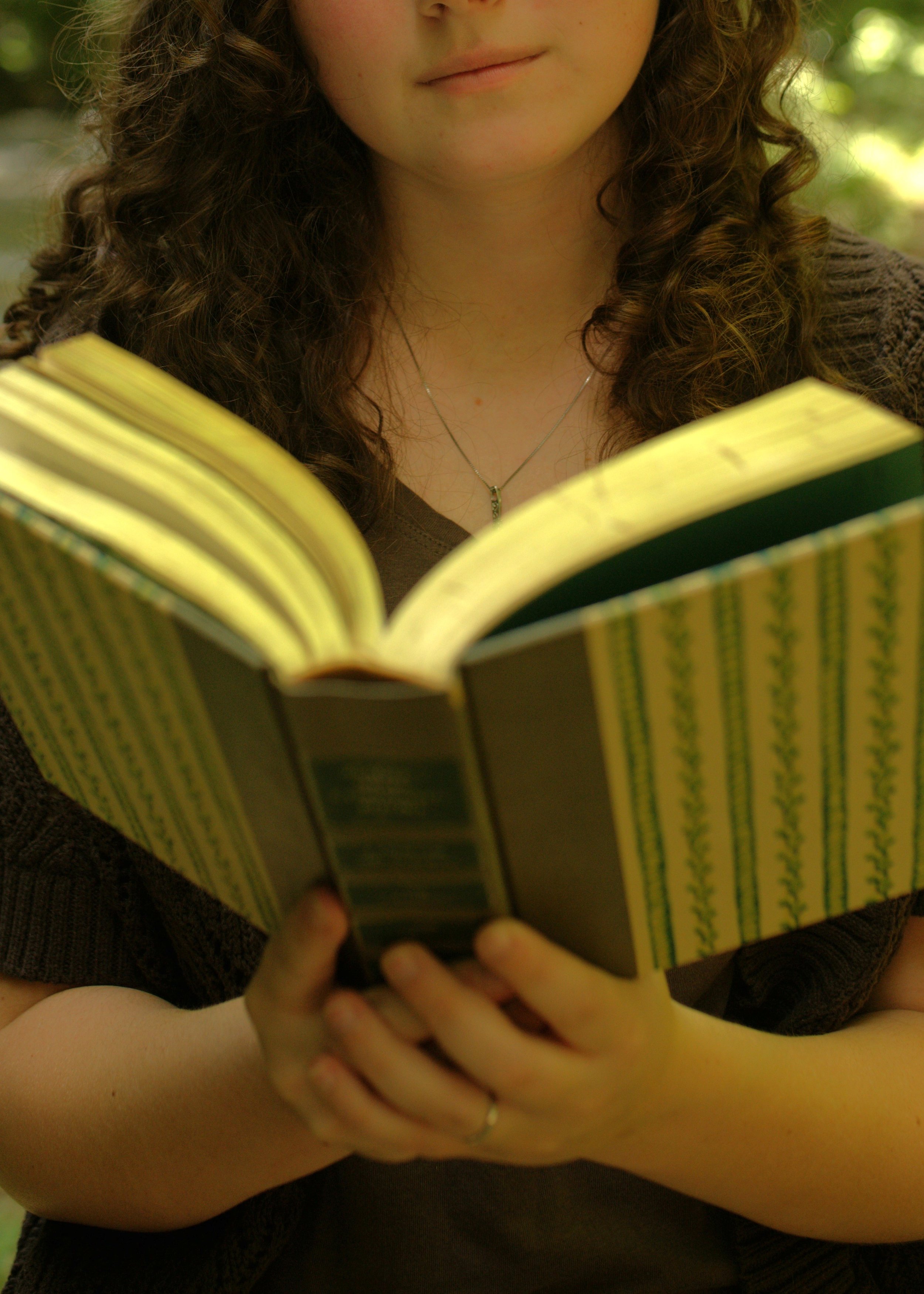 A woman with curly hair is holding an open book and reading outdoors in a natural setting.