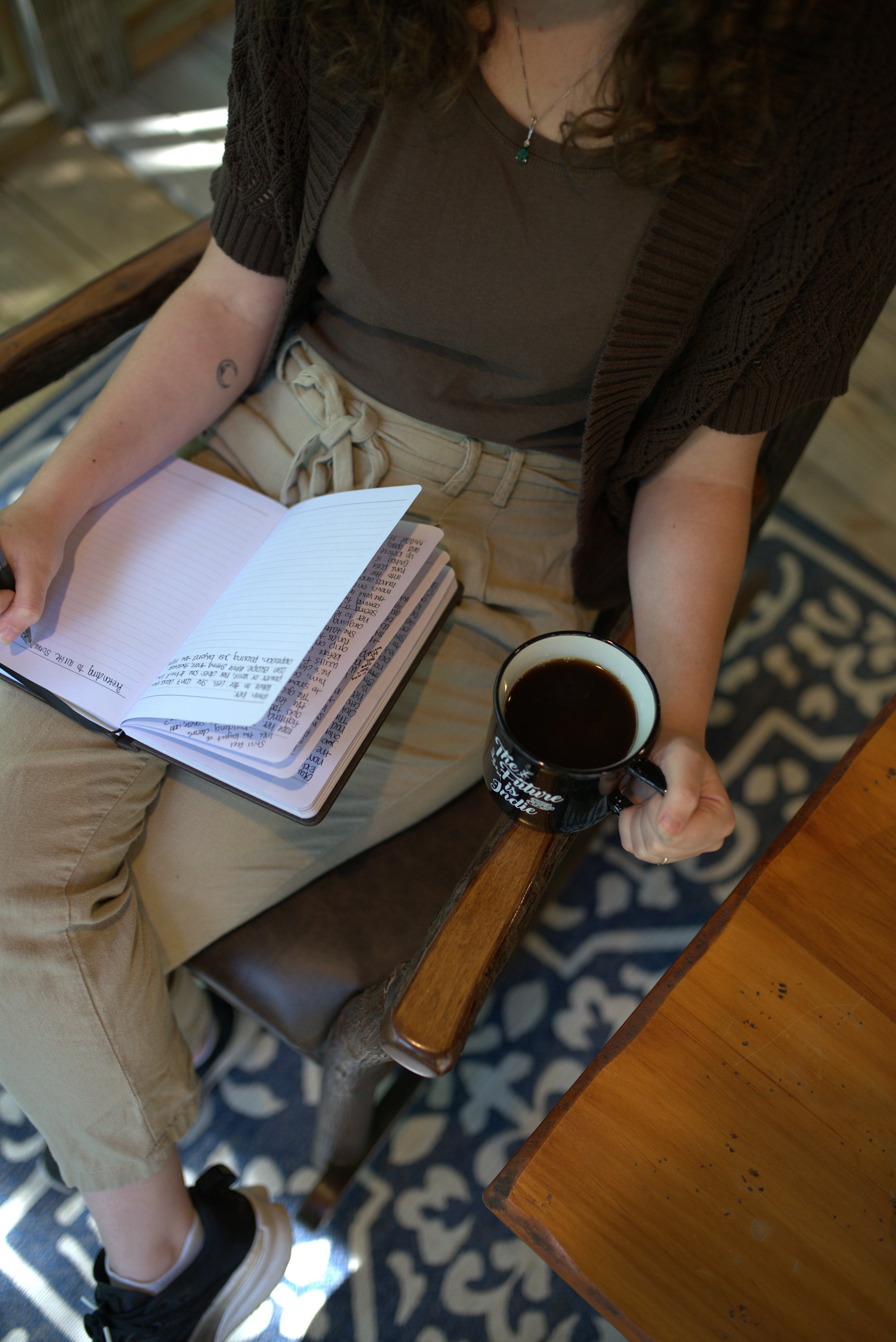 A woman sitting at a wooden table holding a black mug filled with coffee in her right hand and reading a notebook with handwritten notes.