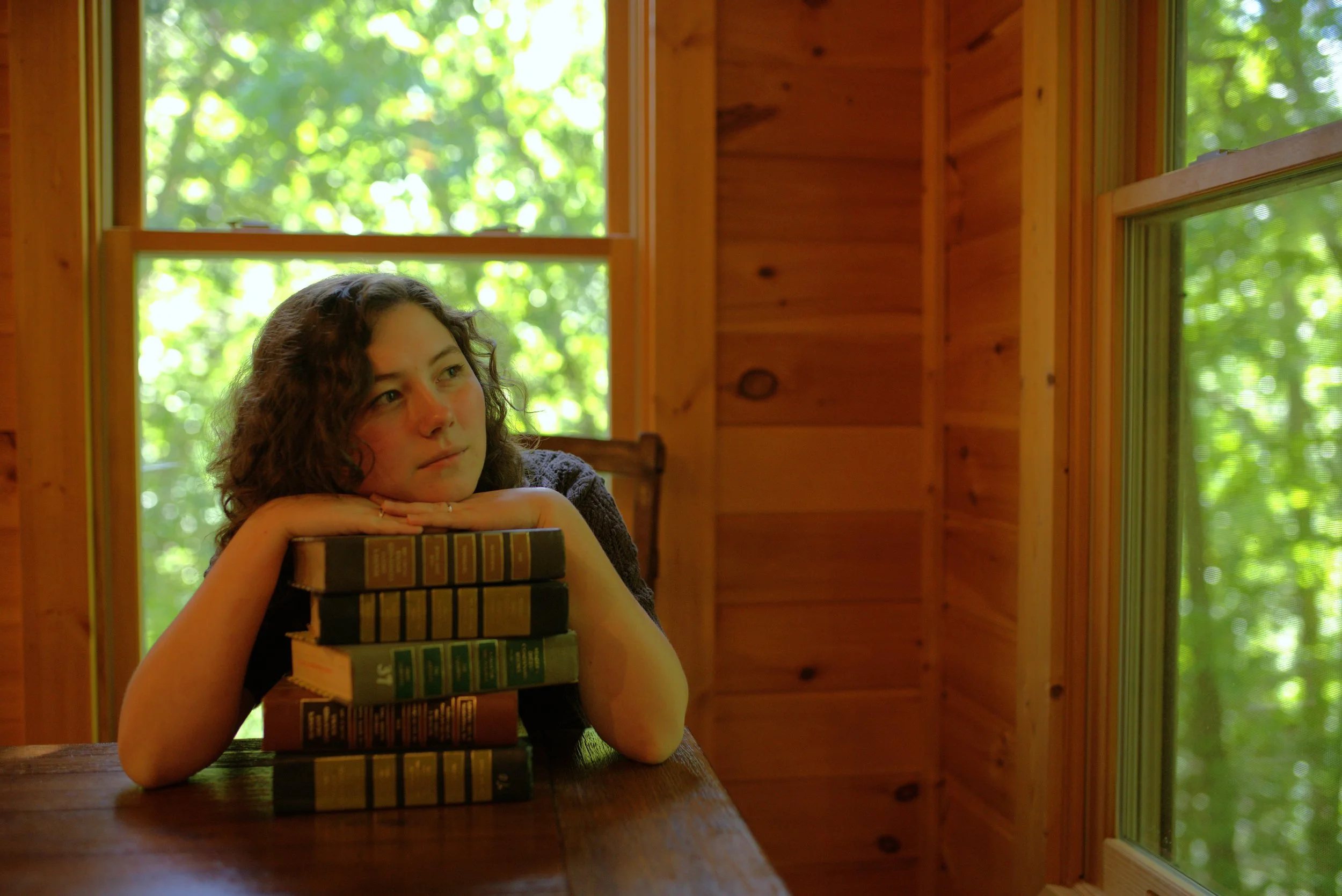 Young woman resting her head on her hands, leaning on a stack of books, inside a wooden cabin with large windows and a view of green trees outside.