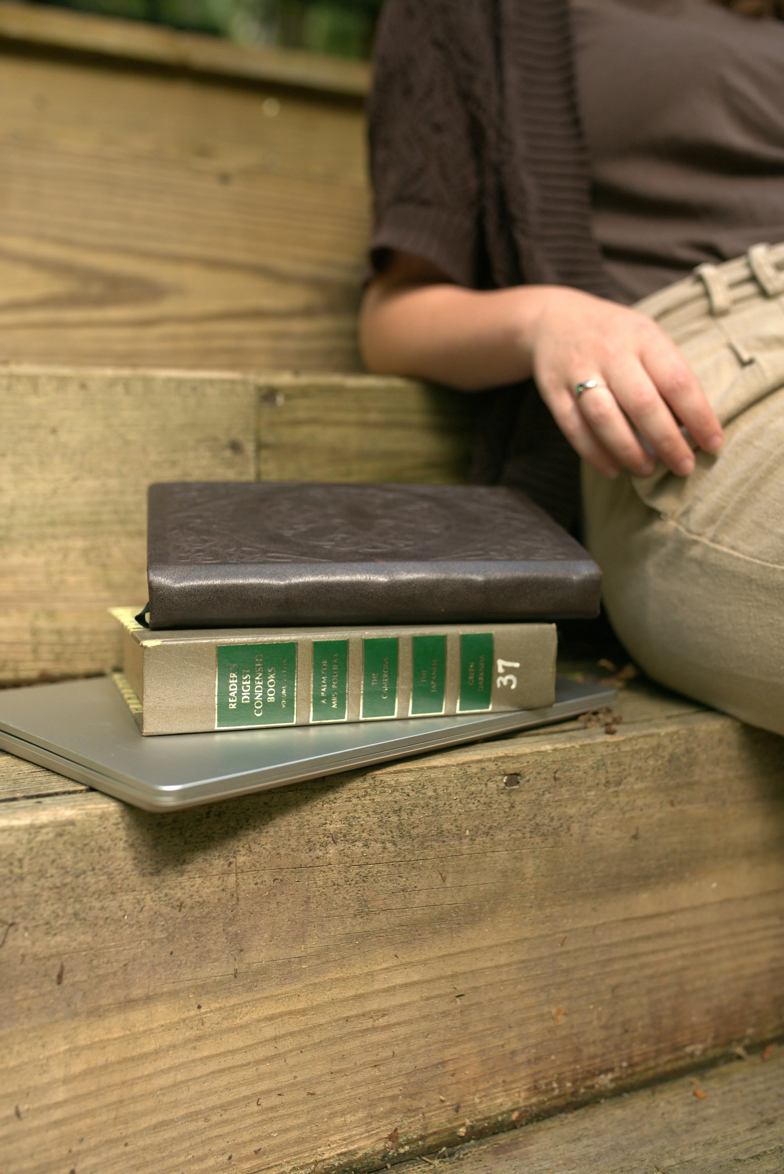 A person sitting on a wooden bench with a gray photo book, a stack of green and black books, and a gray laptop on the bench.