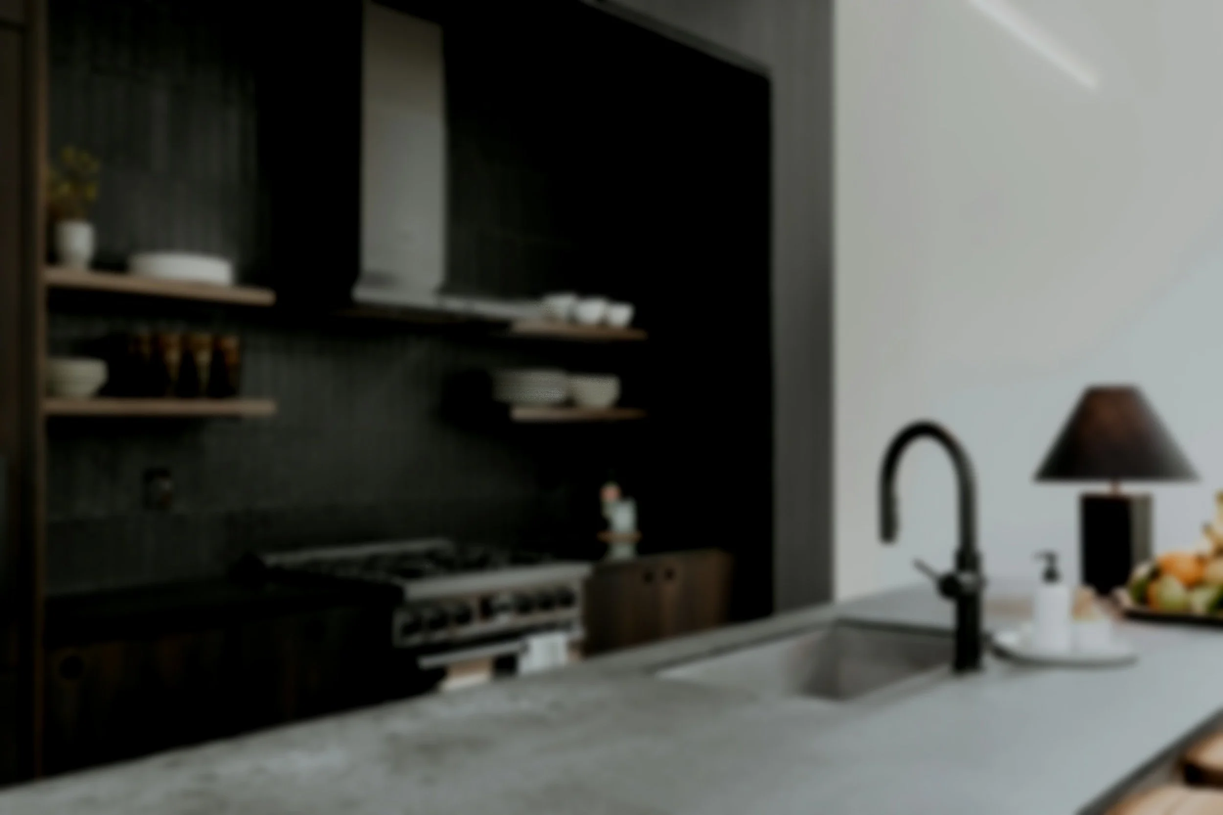 Modern kitchen with black cabinetry, open shelves, a stove, a sink with a black faucet, a black table lamp, and a bowl of fruit on the countertop.