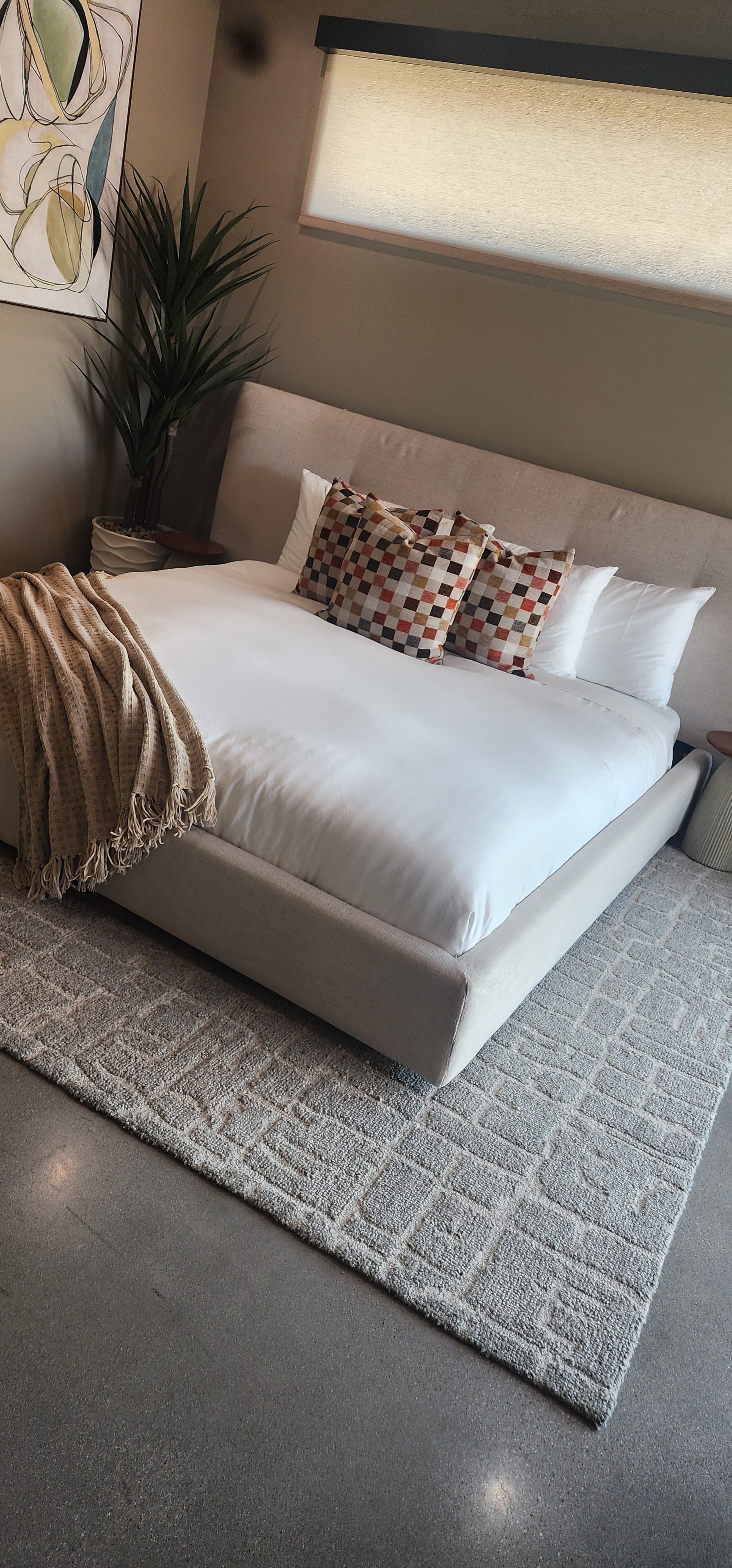 A neatly made bed with white sheets, three striped accent pillows, and a beige throw blanket on a beige upholstered bed frame. There is a potted plant in the corner and a textured rug on the floor, with a window covered by a beige roller shade behind the bed.