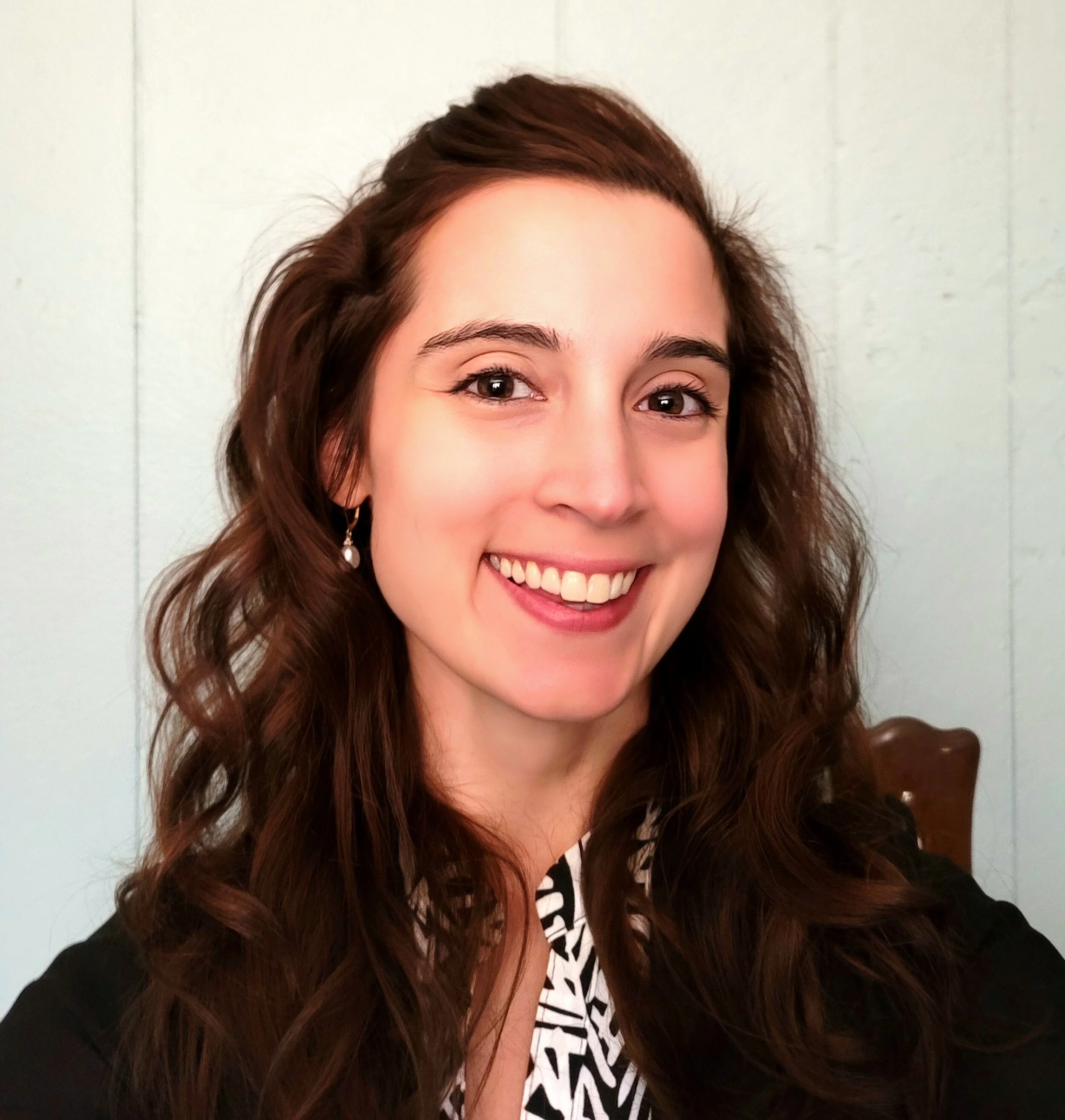 A woman with long, curly brown hair and earrings smiling at the camera, wearing a black jacket over a patterned top, with a light-colored wall in the background.