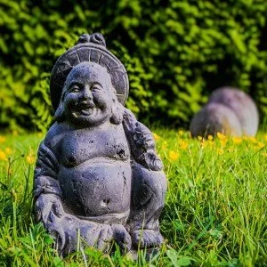 Stone statue of a laughing, chubby Buddha with a joyful expression, sitting in a grassy area with yellow flowers and green foliage in the background.