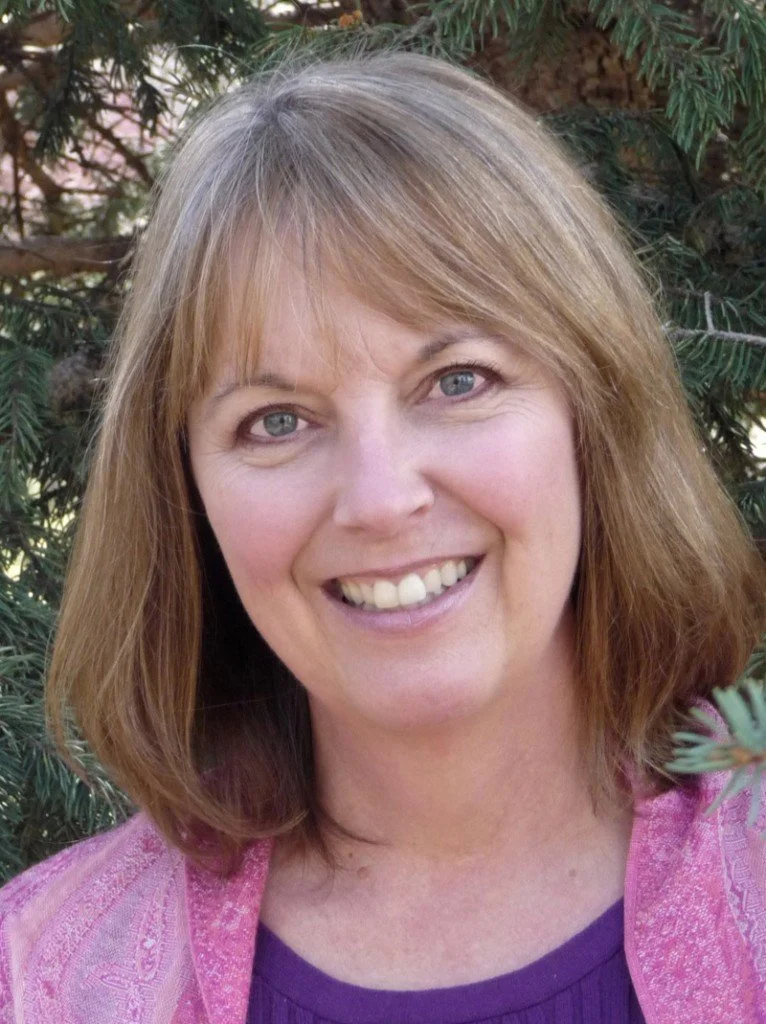A woman with shoulder-length brown hair and blue eyes, smiling, outdoors with green foliage in the background, wearing a purple top and pink shawl.