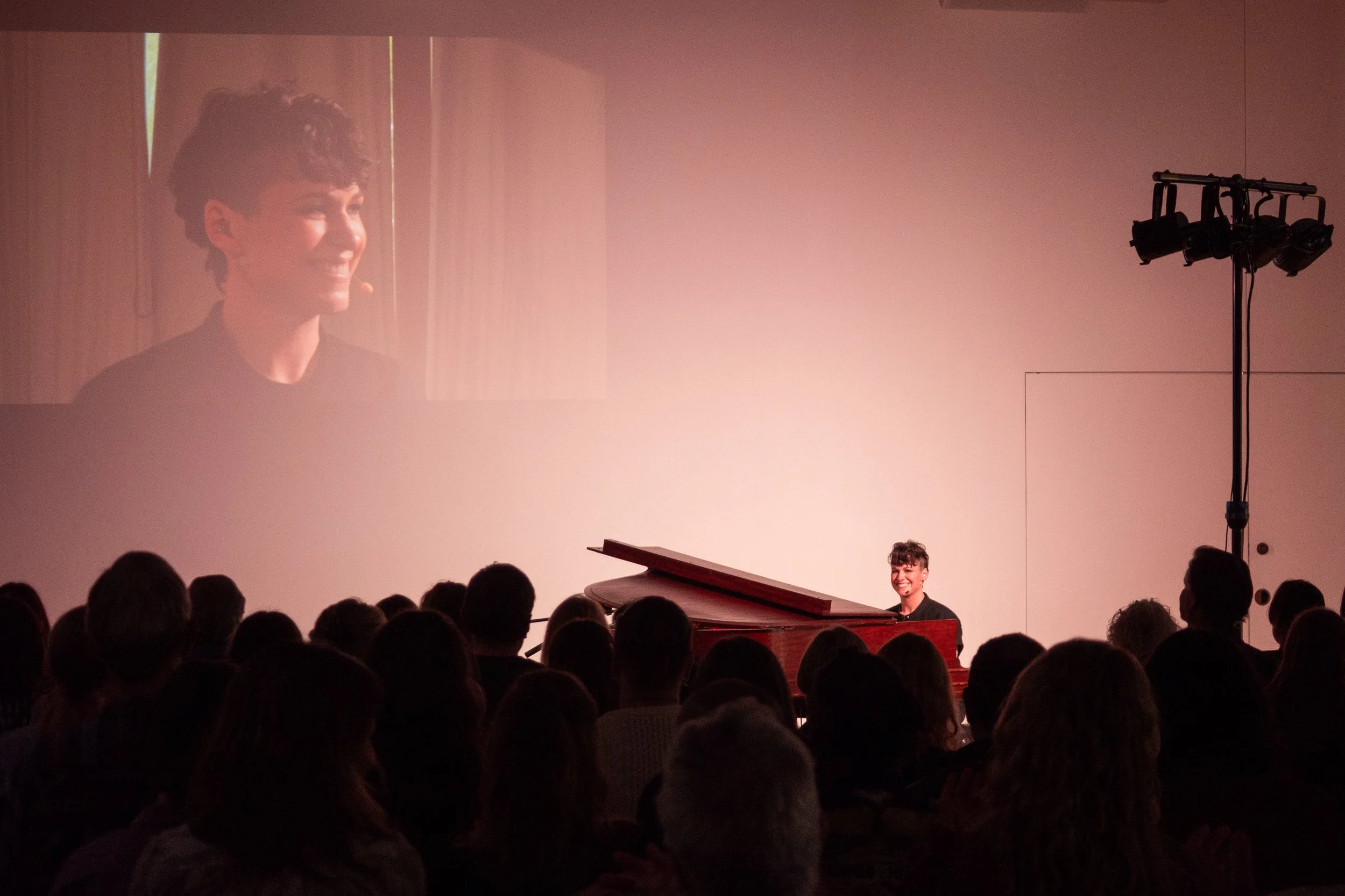 A woman with short curly hair playing a red grand piano on stage, smiling at the audience. A large screen behind her displays her face. Audience members are seated in front of her, and stage lighting and equipment are visible.