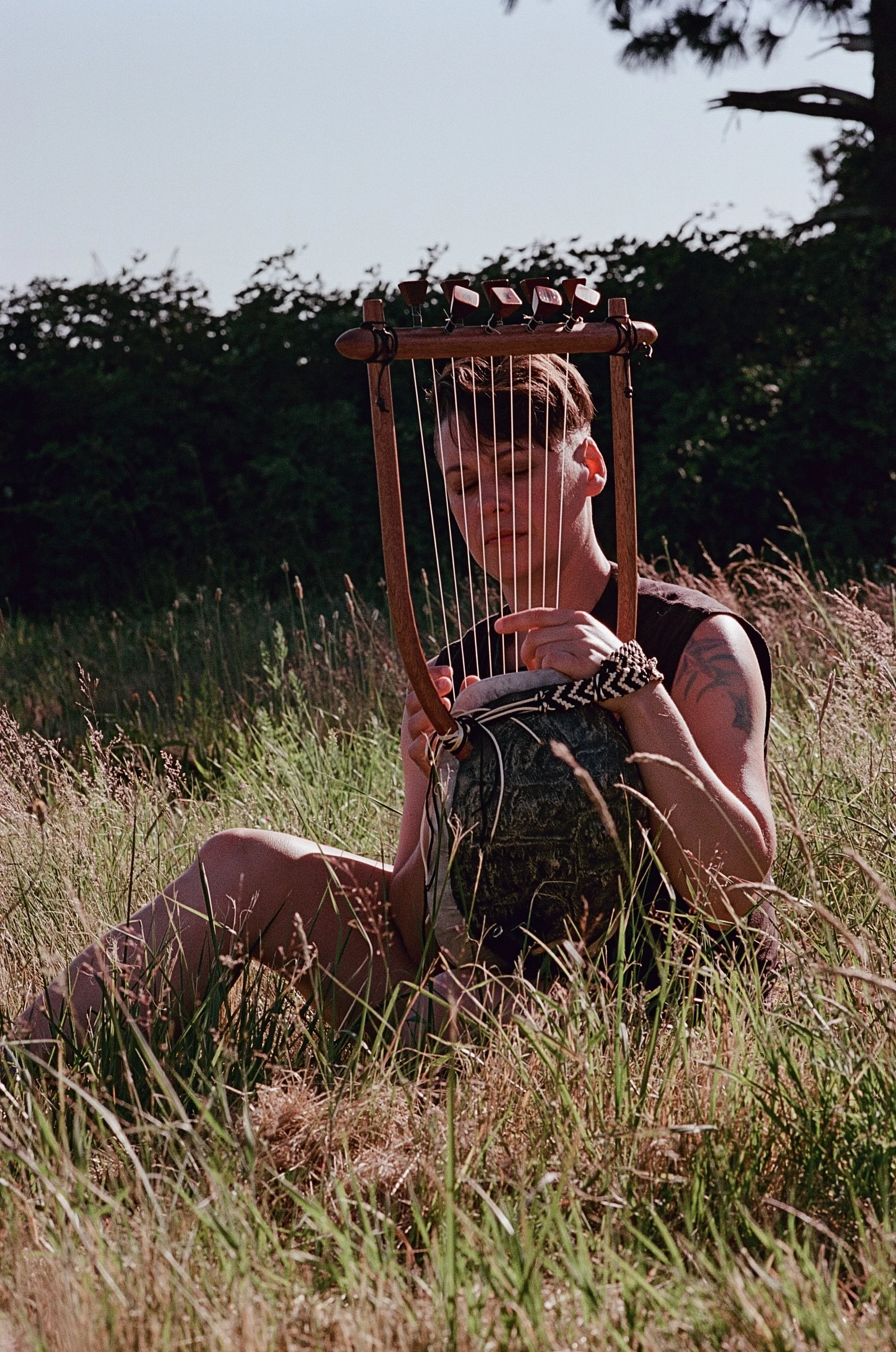 A young person with short hair and a sleeveless shirt sitting in a field of tall grass during daytime, holding a wooden harp close to their face with their eyes closed.