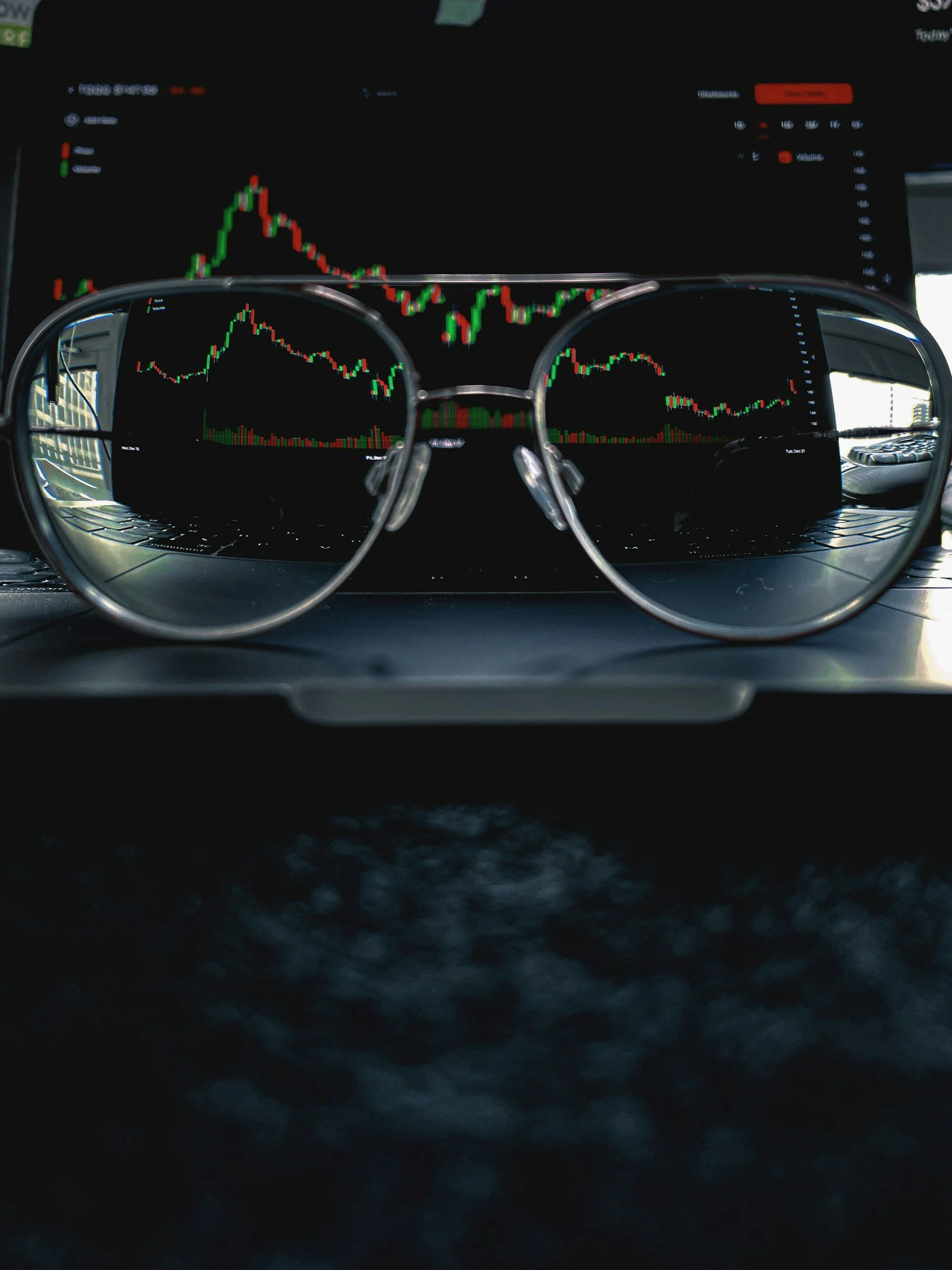 Close-up view of sunglasses with stock market charts reflected in the lenses, placed on a laptop with a larger screen displaying a similar chart in the background.
