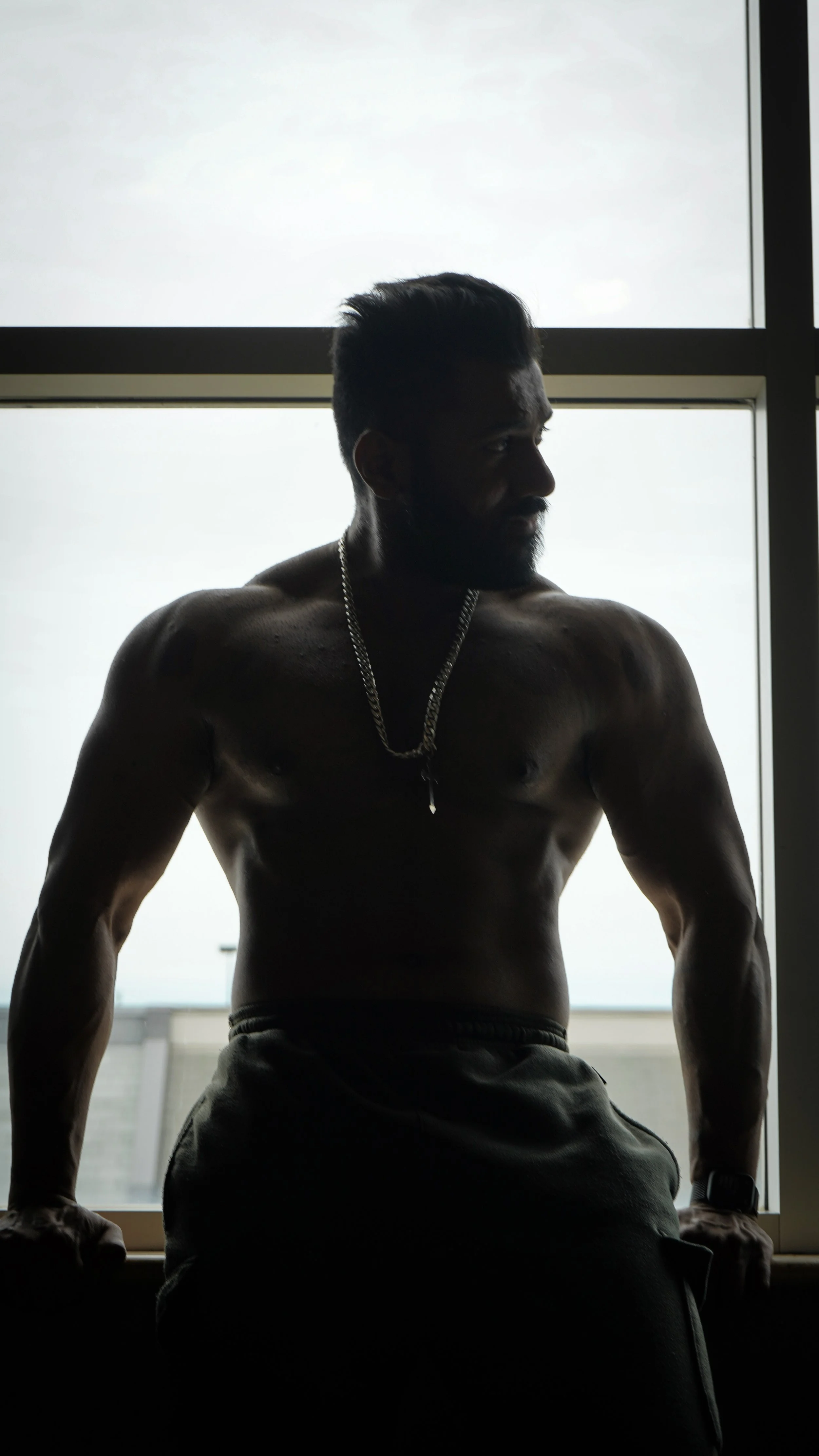 A shirtless man with a beard and short hair wearing a chain necklace and watch, sitting by a window with backlit lighting, casting his face in shadow.