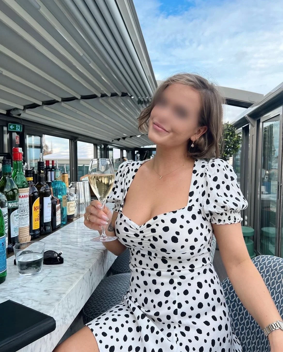 A woman in a black and white polka dot dress holding a glass of white wine, sitting at a bar or restaurant with bottles of alcohol behind her, outdoors with a blue sky.