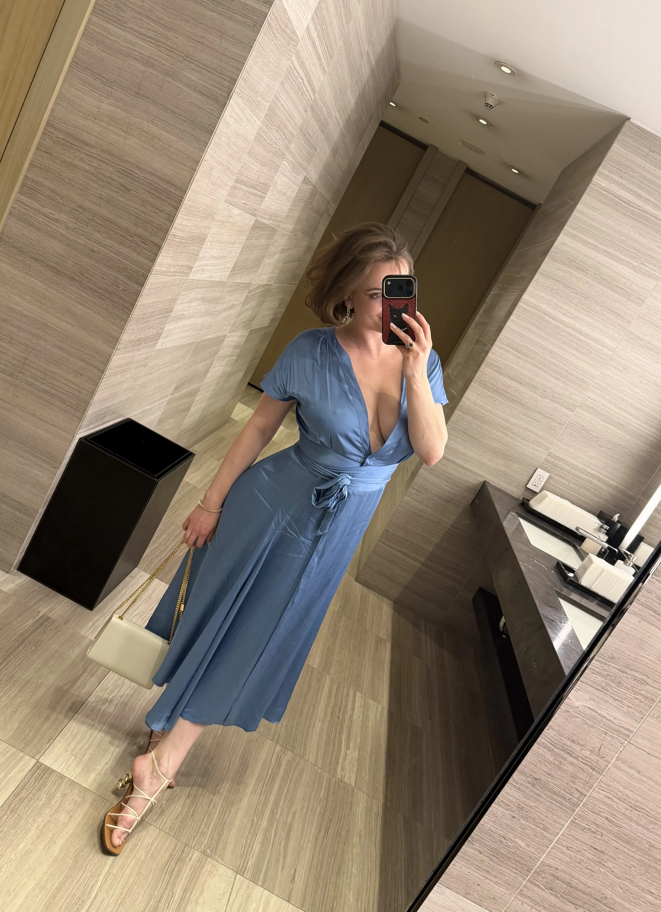 A woman taking a mirror selfie in a bathroom, wearing a blue dress with a deep neckline, beige high heels, and holding a white handbag with a gold chain. The bathroom has beige tiles and a black countertop.
