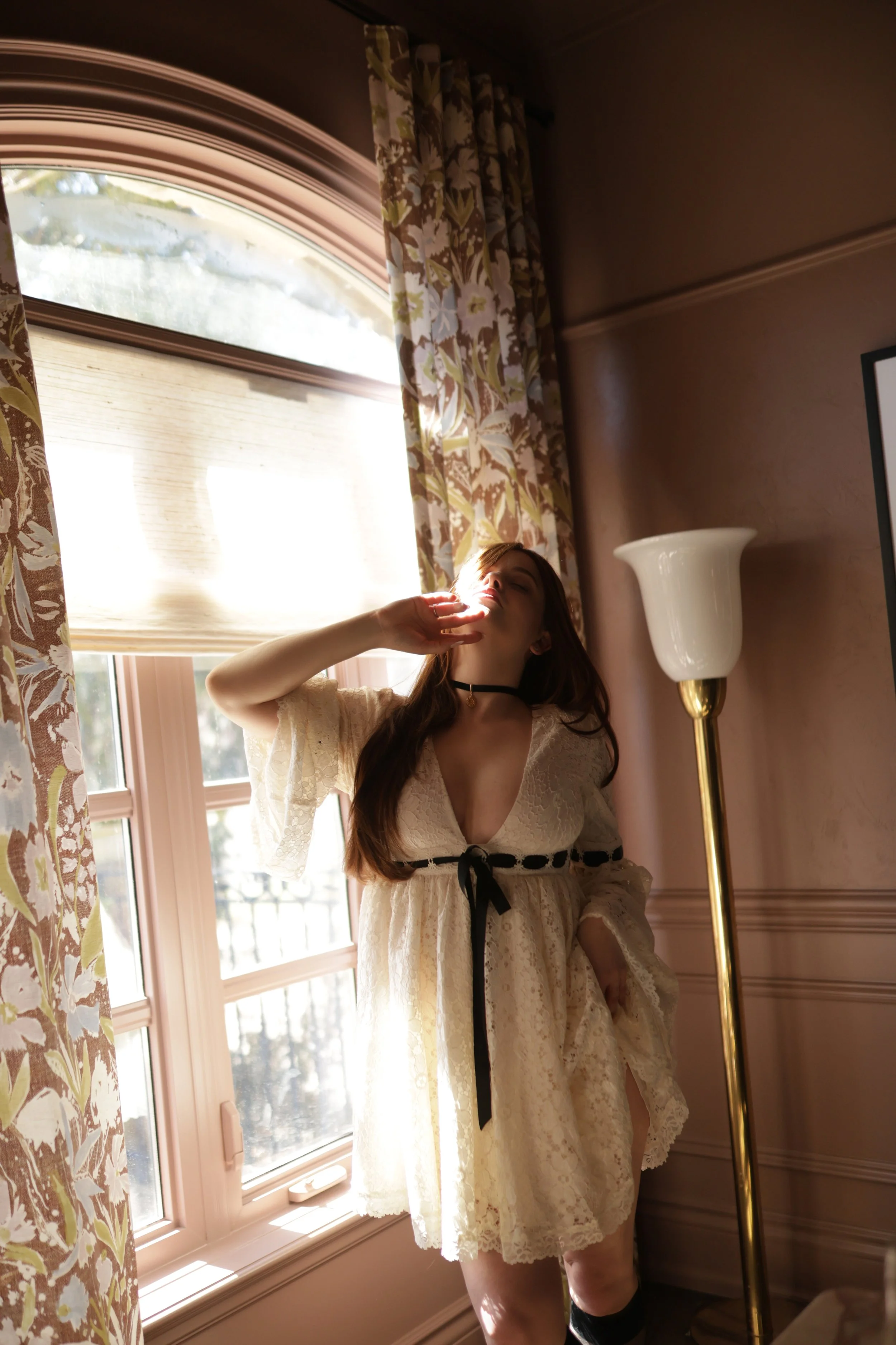 Woman with red hair wearing a white dress with black ribbon standing next to a window