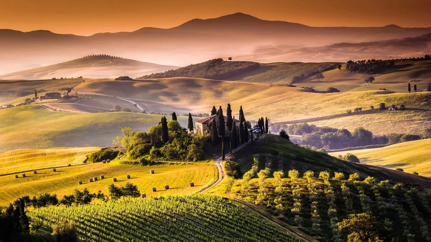 Scenic landscape of rolling hills with farm fields, scattered trees, and a distant mountain range during sunset, featuring a path leading to a cluster of buildings surrounded by tall cypress trees.