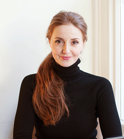 A woman with long red hair wearing a black turtleneck, sitting in front of a window.