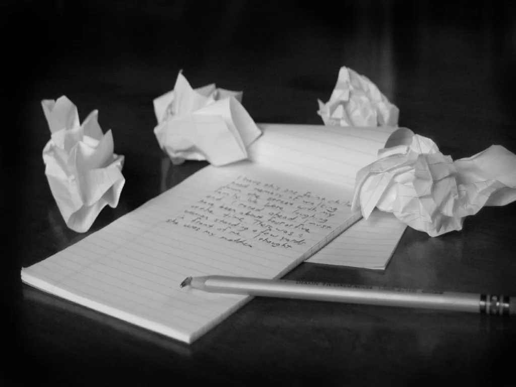 A notebook with handwritten text on a table, surrounded by crumpled pieces of paper and a pen.