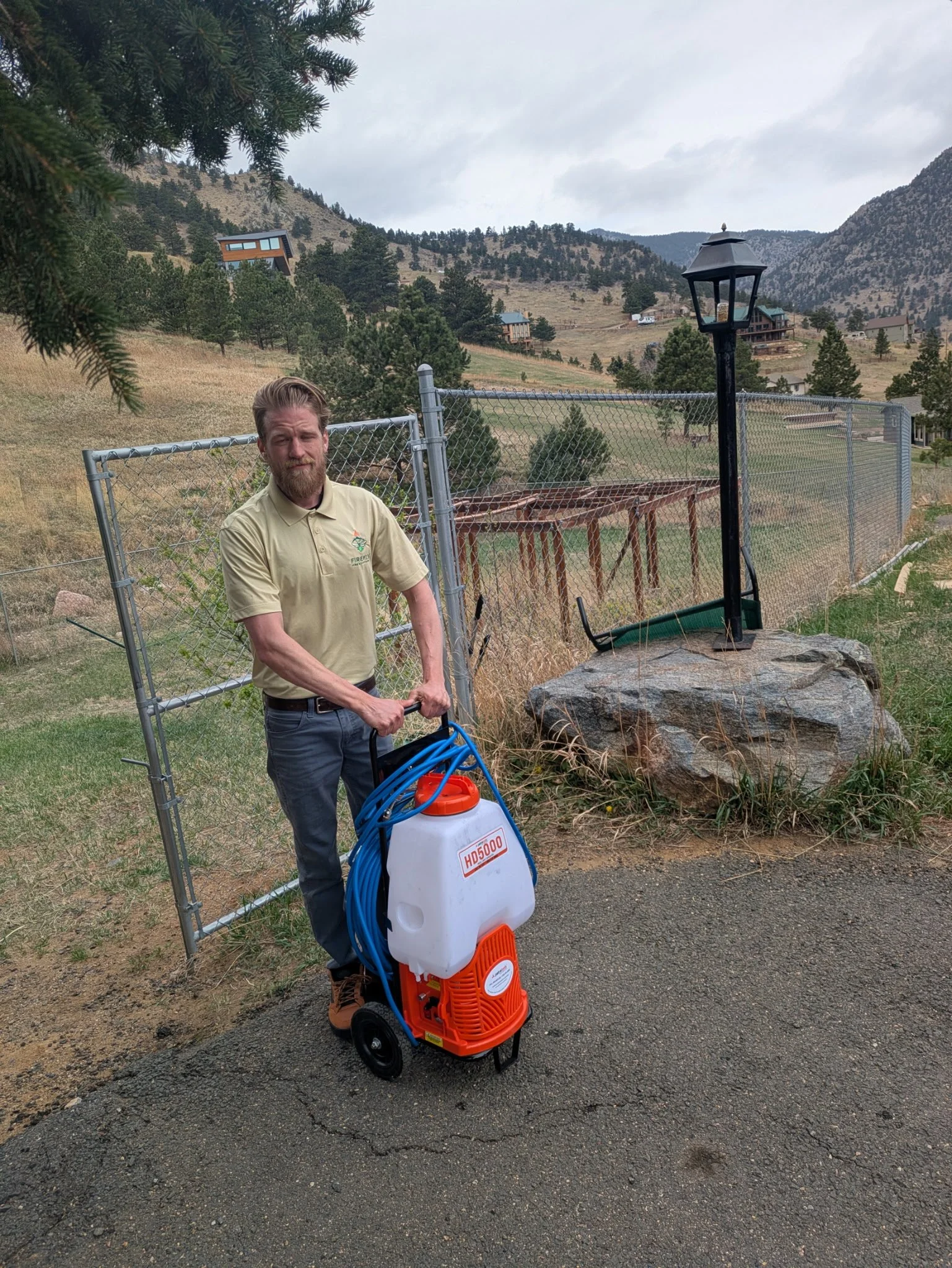 Man with beard wearing beige polo shirt standing outdoors on paved path, holding a portable sprayer with a large white tank, orange base, and blue hose, against rural hillside background with fences, houses, and trees.