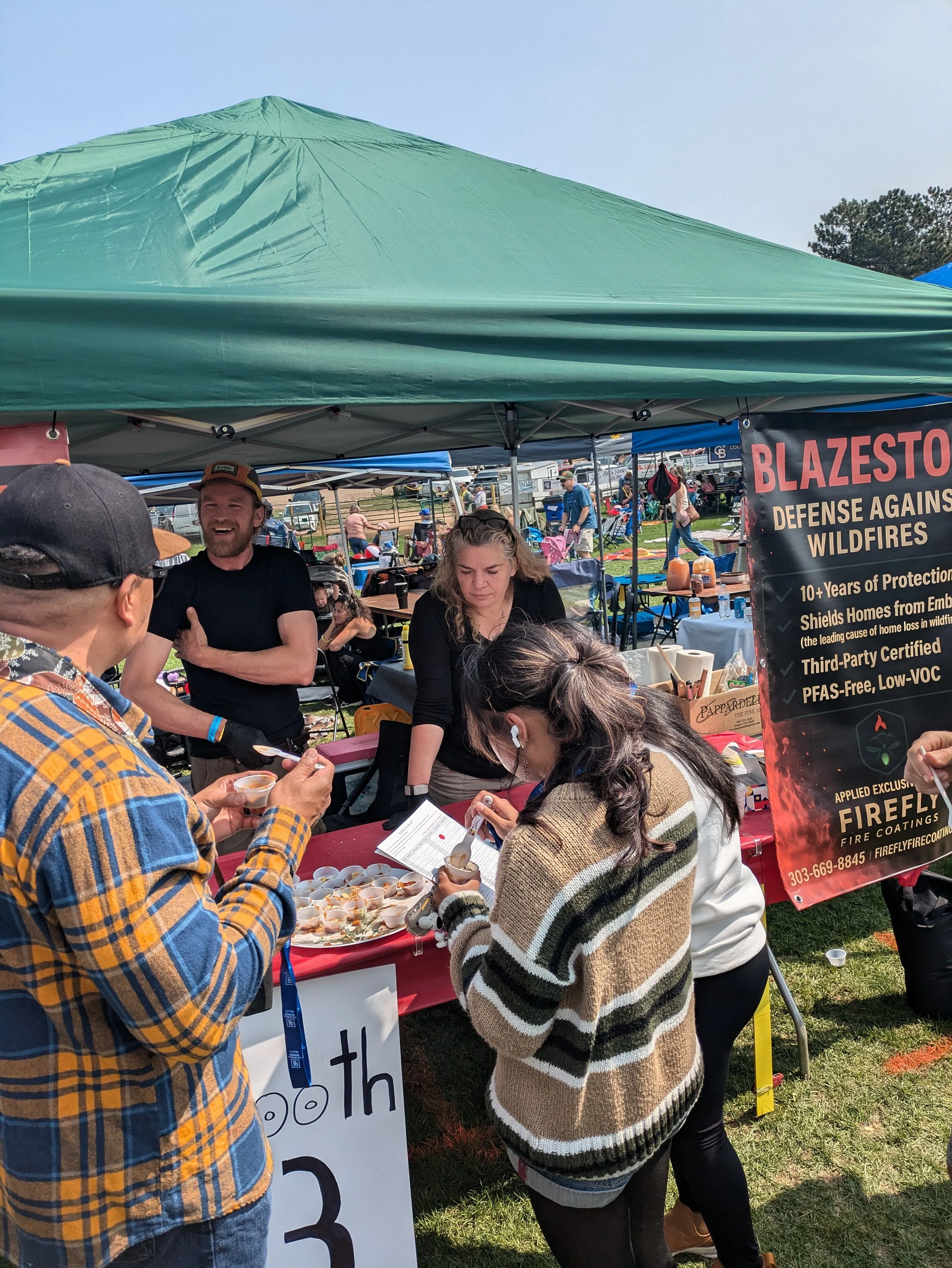 People at an outdoor booth at a fair or festival, with a woman serving samples and others engaging, under a green canopy. There's a sign about wildfire protection products.