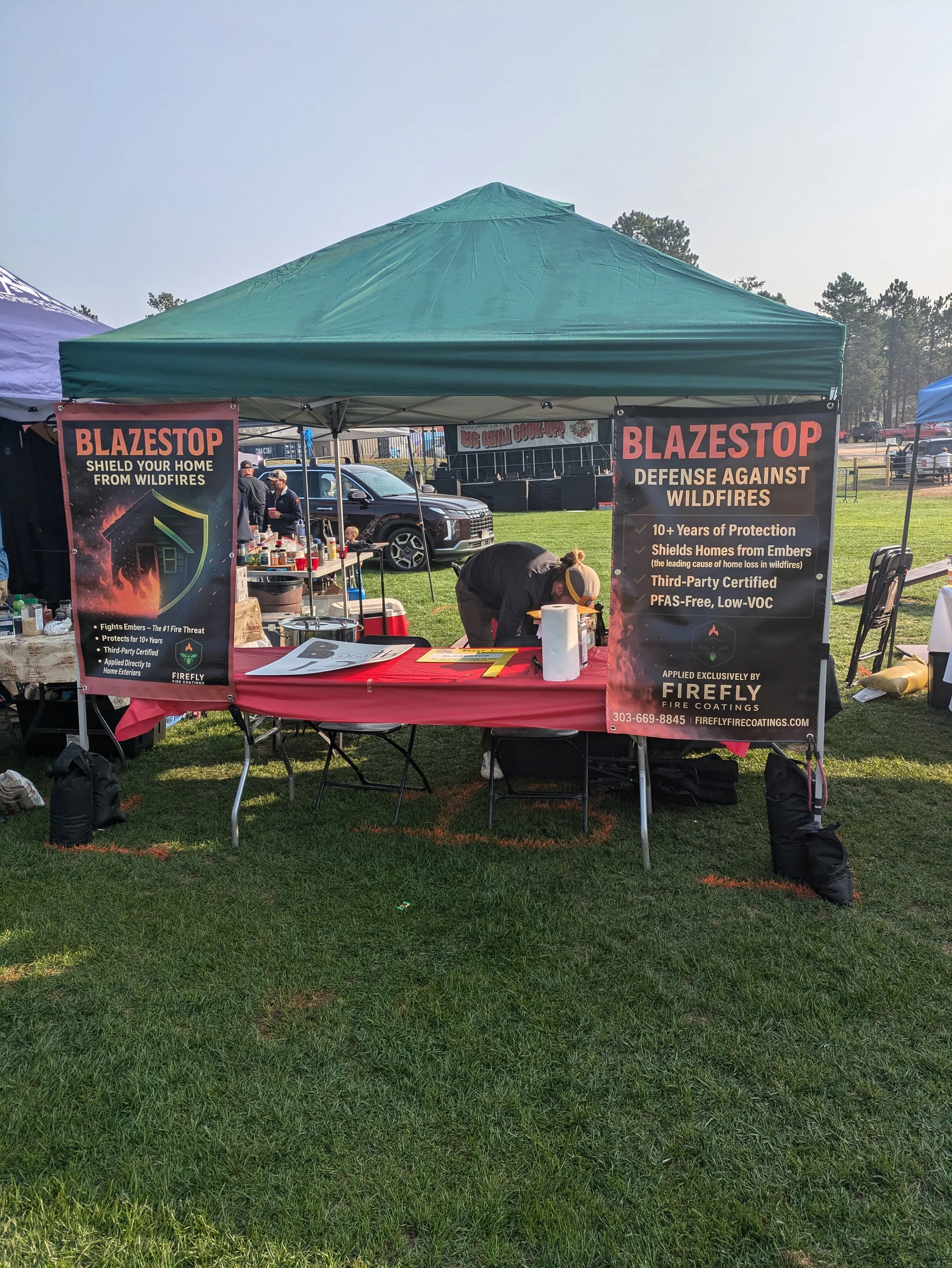 A green canopy tent with two large red and black banners promoting fireproof home coatings, set up on a grassy outdoor area at a fair or event, with a person bending over a table underneath.
