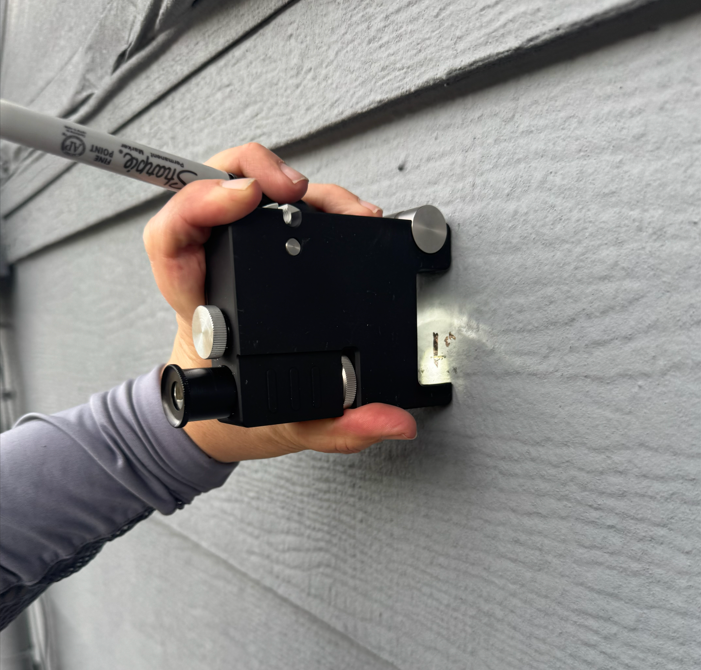 A person installing a black streak camera on a gray exterior wall using a marker to mark the placement.