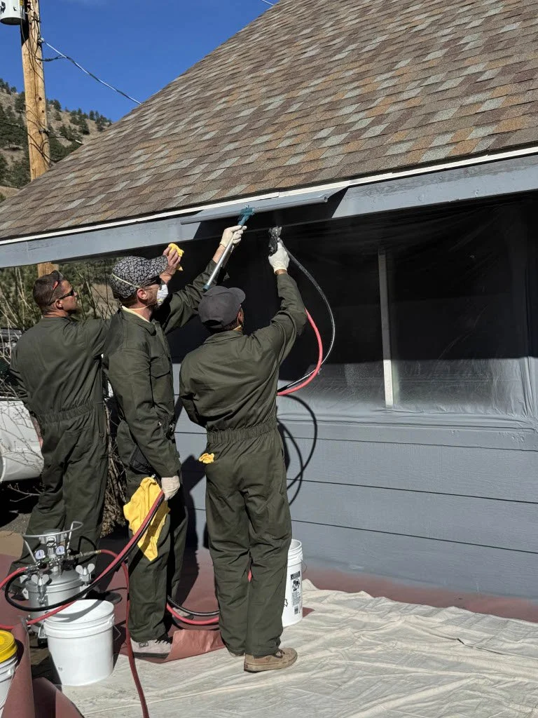 Three workers in protective suits are spray painting the gutter of a house's exterior wall grey, with one person holding a spray gun, one holding a yellow cloth, and the third observing. They are outdoors on a sunny day.