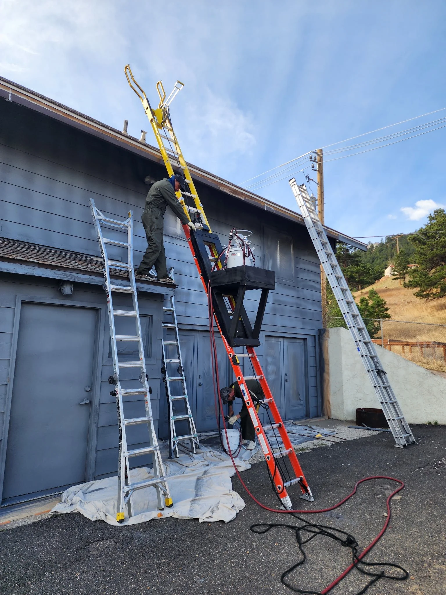 Two workers are on ladders fixing a power line on the side of a building. The scene is set outdoors during the day, with blue sky and some clouds.
