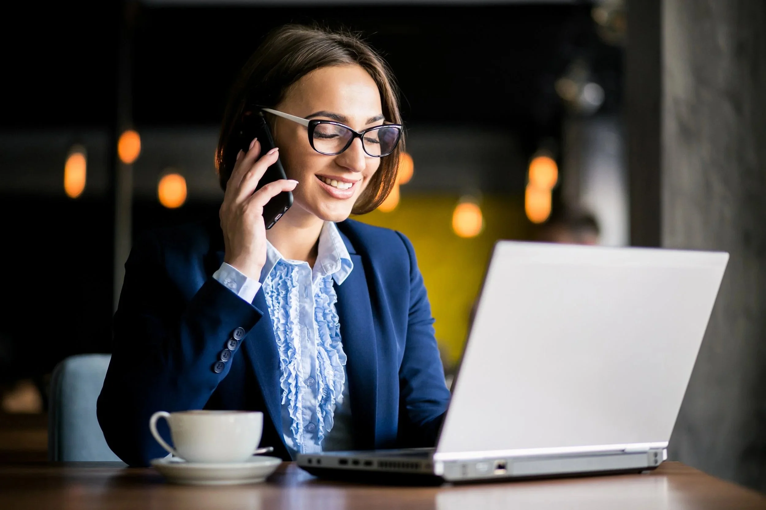 A smiling woman in business attire, wearing glasses, sitting at a table with a laptop and a cup, talking on her cellphone in a modern cafe or office setting.