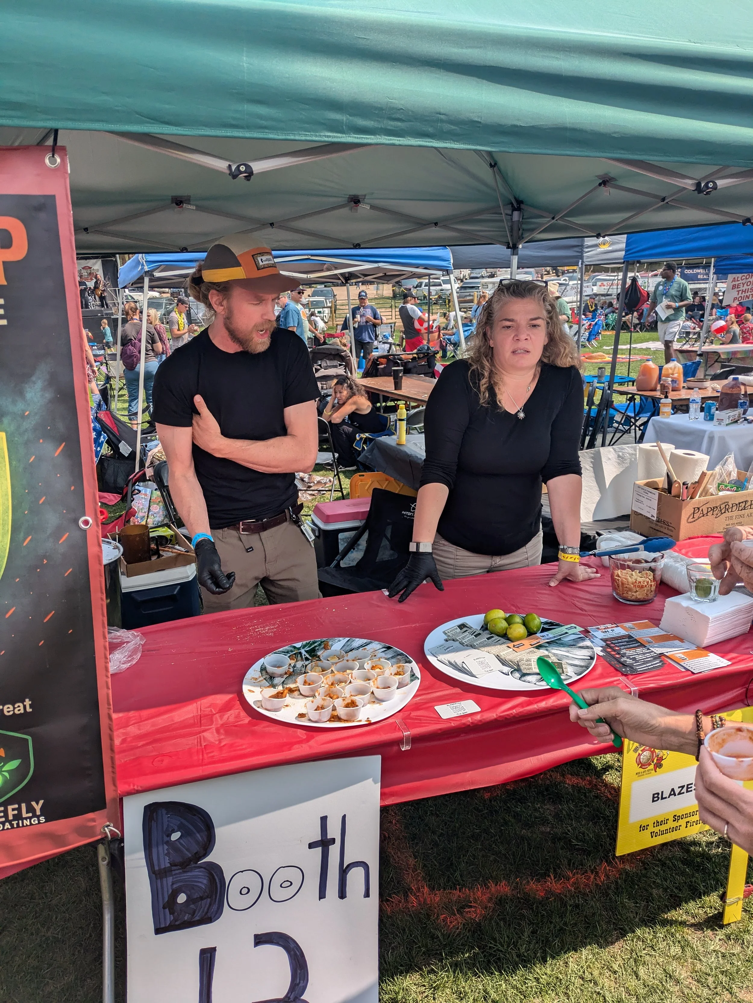 People standing behind a red table at an outdoor event, with one woman looking serious and a man wearing a cap and gloves. There are limes, small cups of food, and promotional materials on the table.