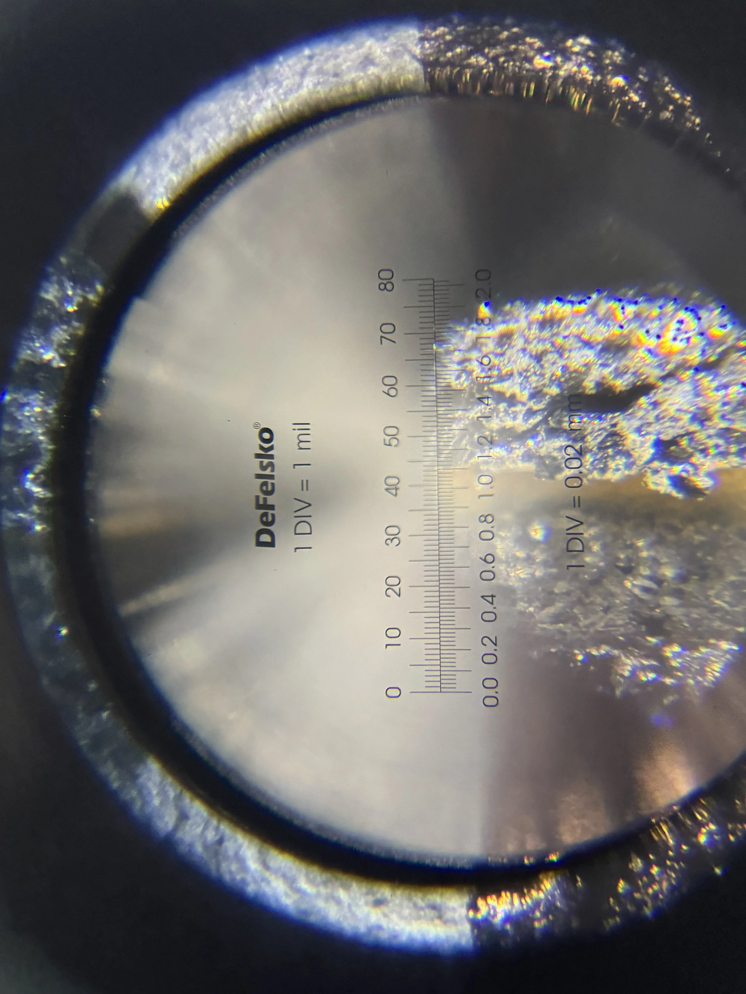 Close-up of a metallic surface through a magnifying glass with measurement markings, showing a textured area on the right side.